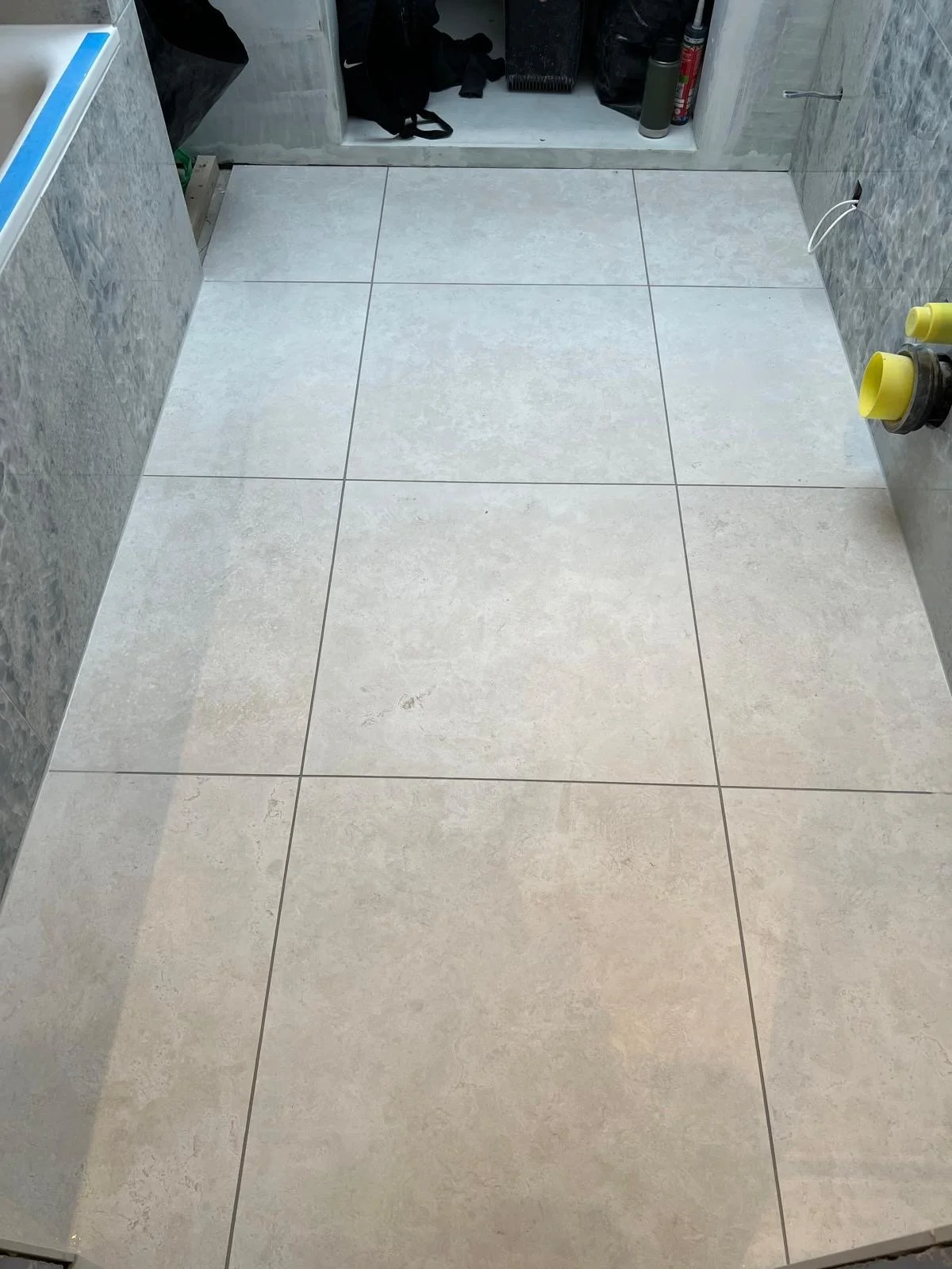 Freshly installed cream-colored ceramic tiles on the floor of a bathroom or utility space, with a small storage area in the background containing cleaning supplies and a black bag.