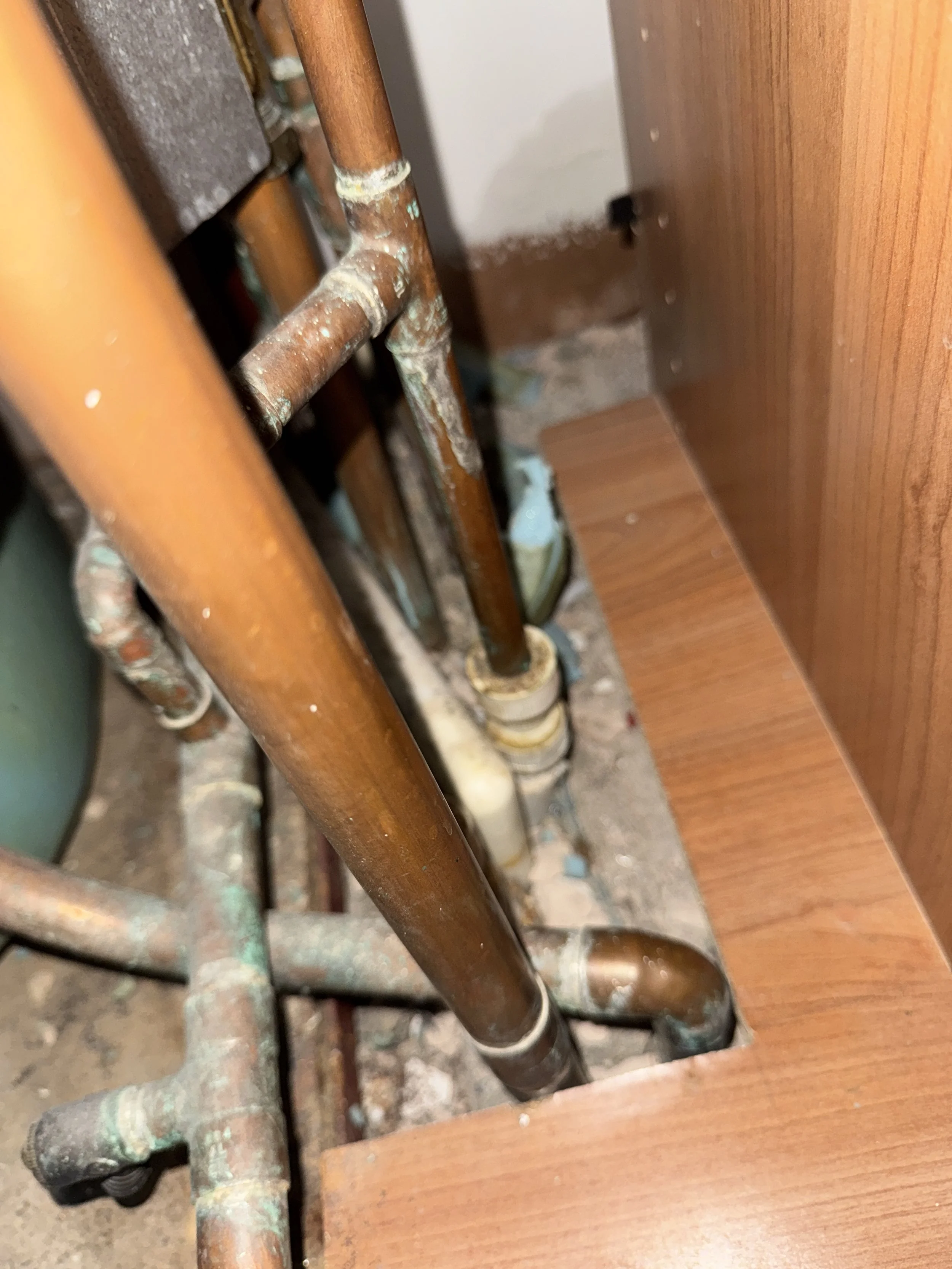 Close-up of a patchy, rusty copper pipe connected to a valve or fitting, next to a wooden surface.