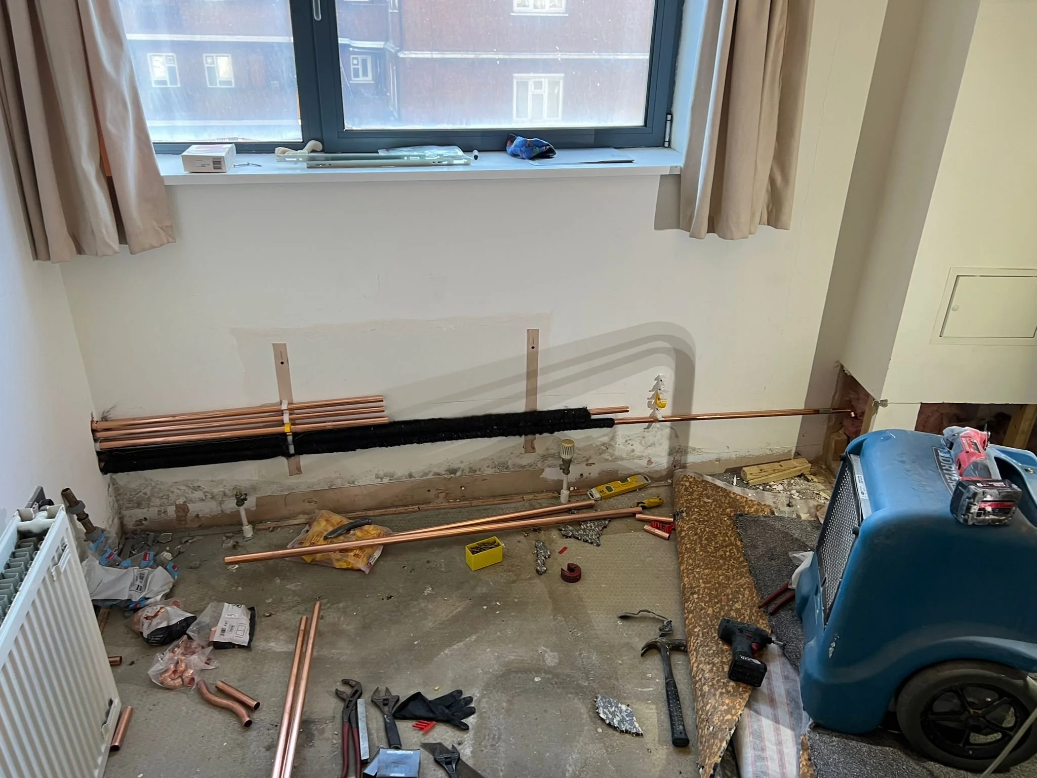 A room under renovation with exposed copper pipes along the bottom of the wall, construction tools and supplies scattered on the floor, a plastic blue container, and a window with beige curtains.