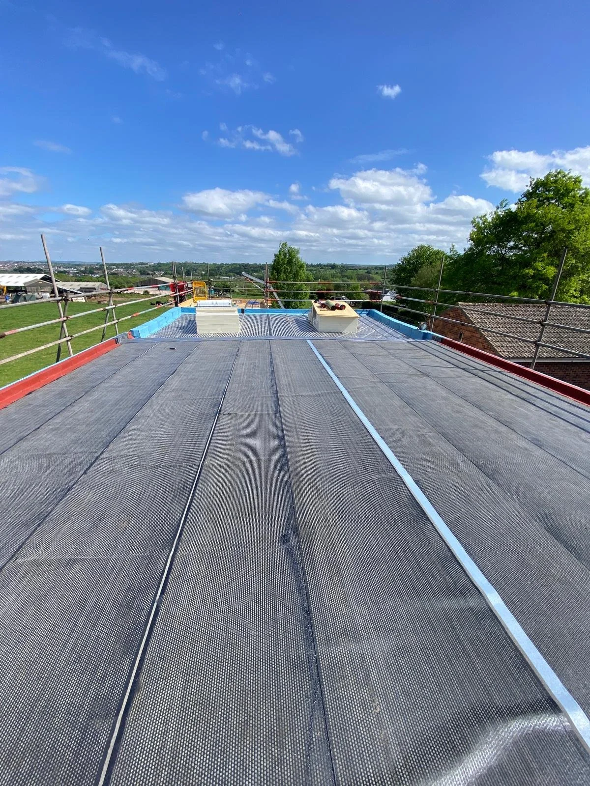 View of a building rooftop with construction safety railing, blue sky with scattered clouds, and distant greenery and rooftops.