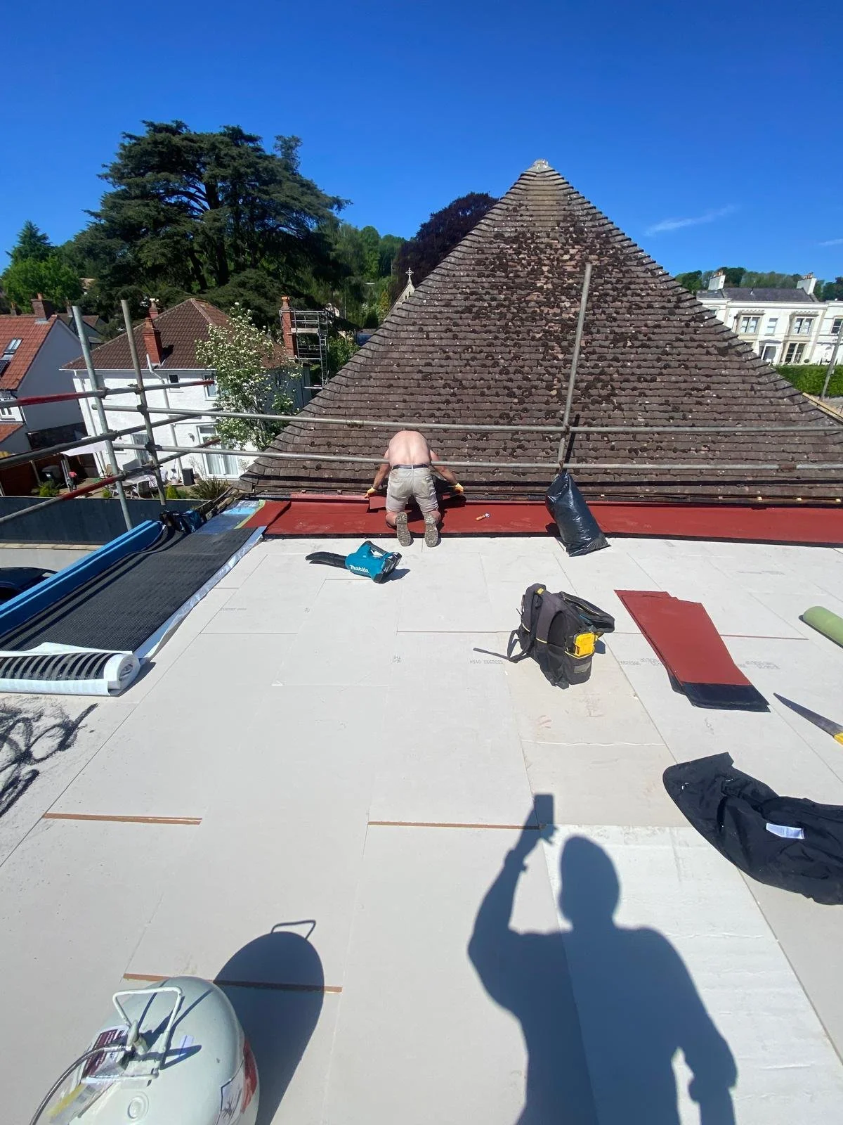 A construction worker on a roof, installing red roofing material near a large, sloped tiled roof, with various tools and supplies around him, under a clear blue sky.