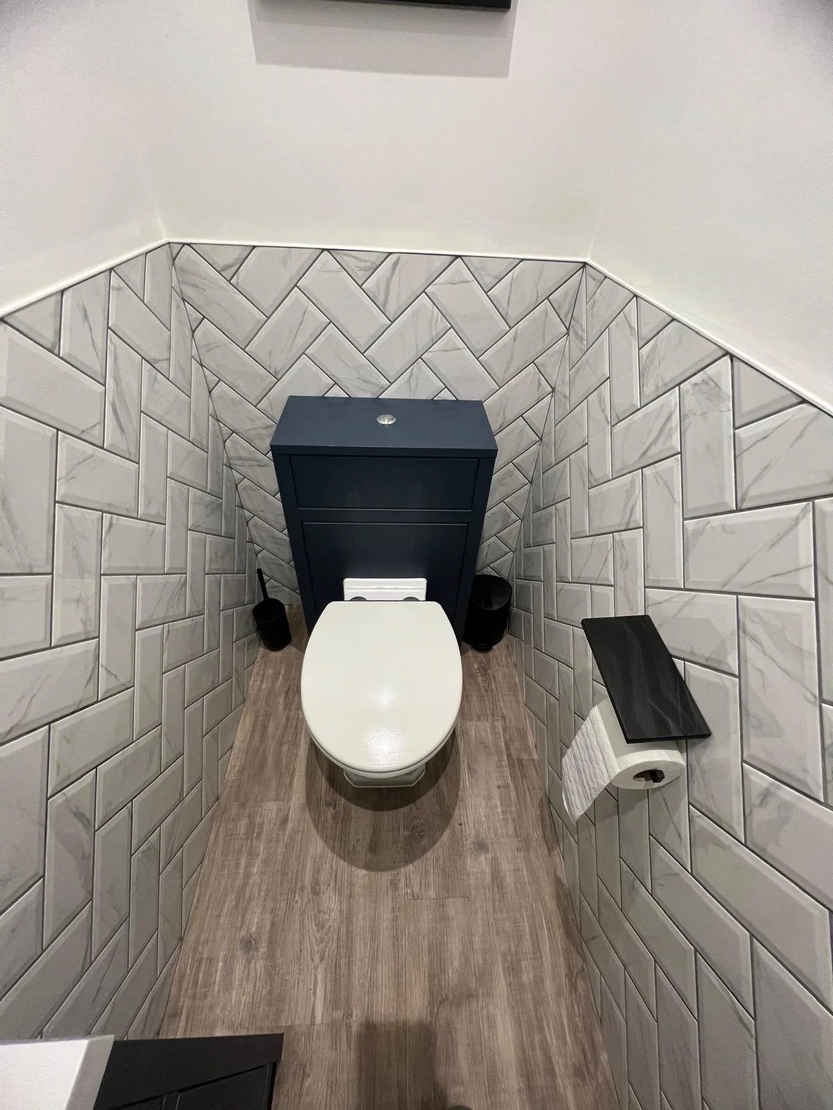 Small bathroom with a white toilet, black toilet brush and holder, and a black rectangular shelf with toilet paper, enclosed by beige and white marble tiles on the walls and wood-colored vinyl flooring.