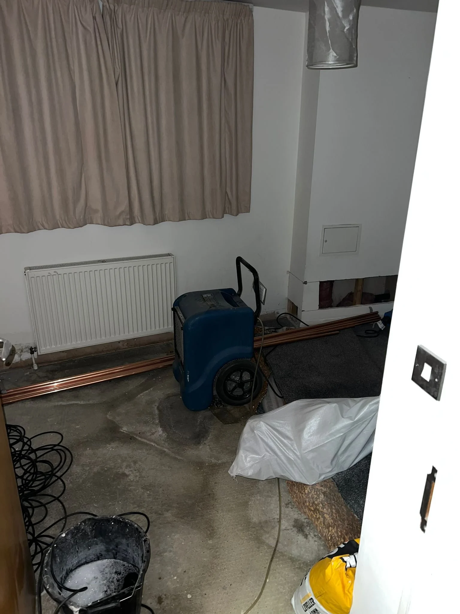 A partially renovated room with a beige curtain covering a window, a white radiator below the window, a black and blue portable heater, a white plastic bag, a yellow bag of instant oatmeal, a gray and black carpet, some wooden molding, some electrica