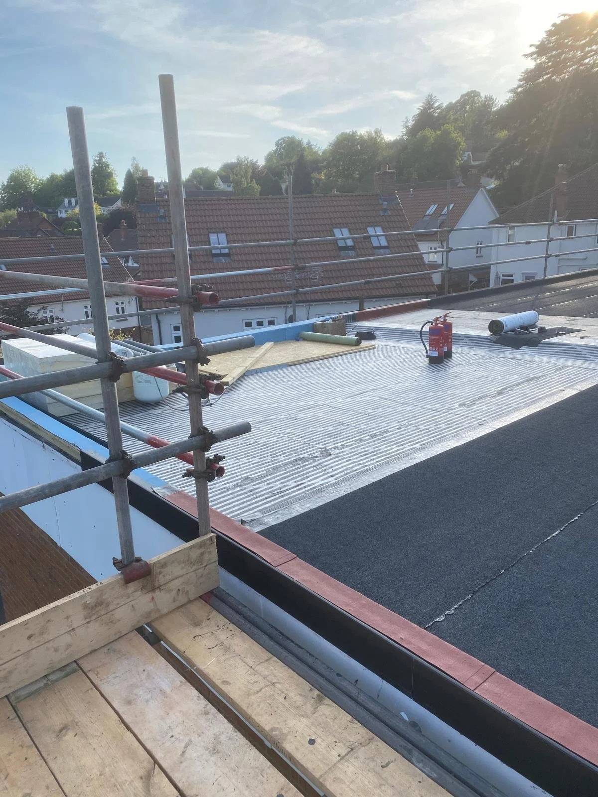 Construction site on a rooftop with scaffolding, roofing materials, and tools, overlooking neighborhood houses with trees under a partly cloudy sky.