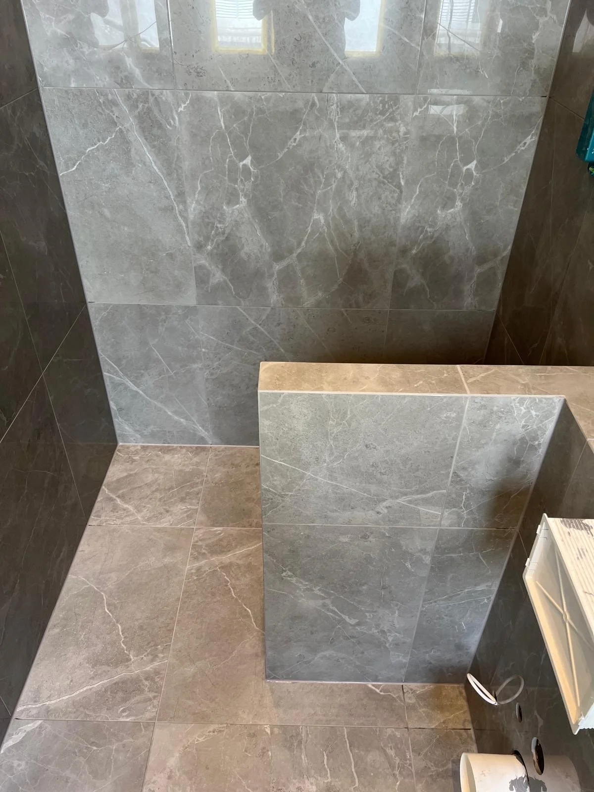 A bathroom with gray marble tiles on the walls and beige tiles on the floor. There is a partial view of a white heated towel rack and a small white container.