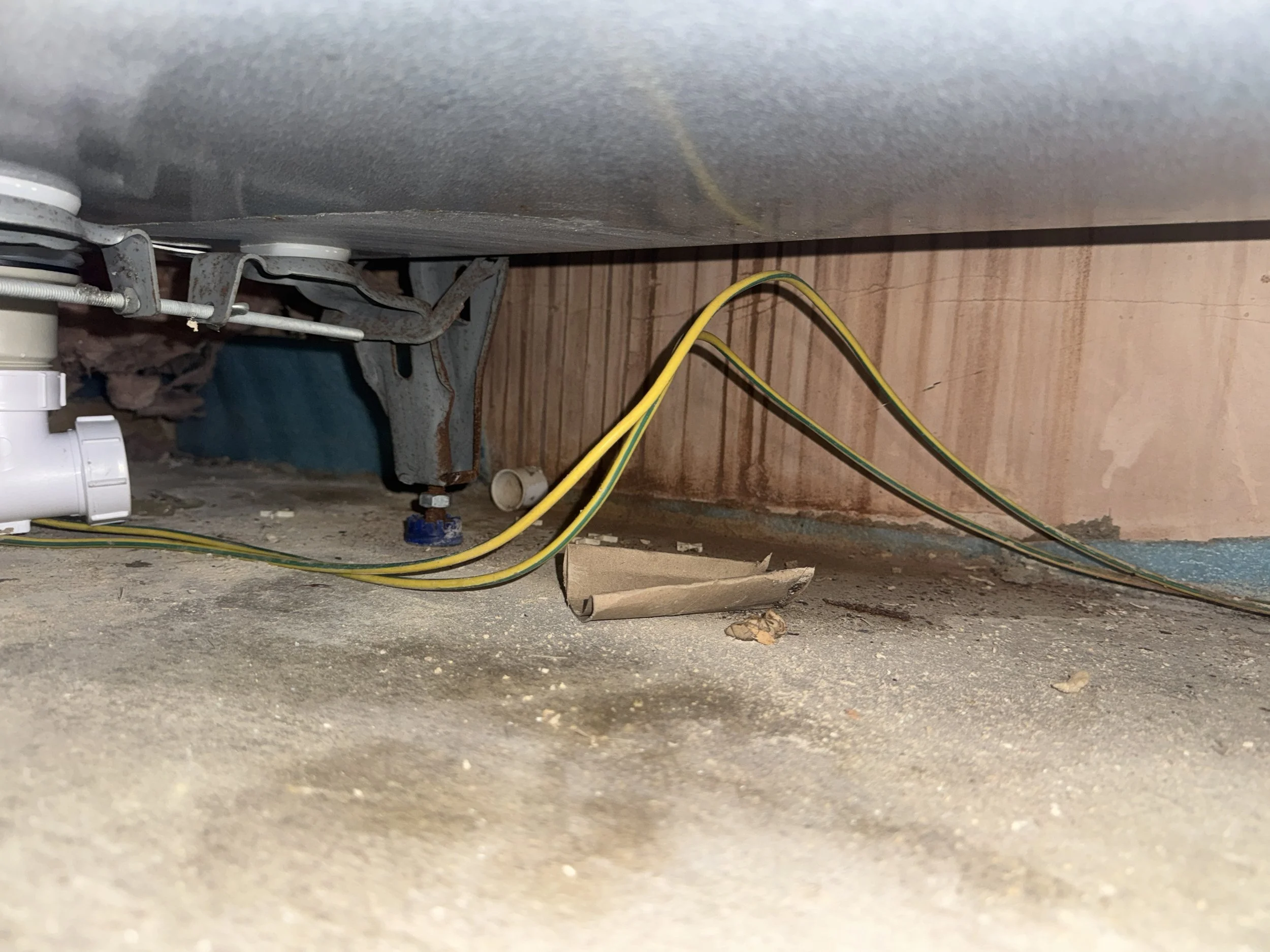 Underneath a kitchen sink showing the pipes, plumbing, and electrical wires attached to the wall and floor.