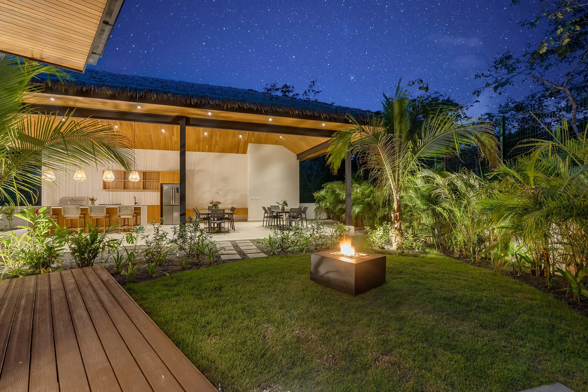 Night view of an outdoor patio with a fire feature, surrounded by lush tropical plants and trees, an open kitchen area with a wooden ceiling, pendant lights, and dining tables.