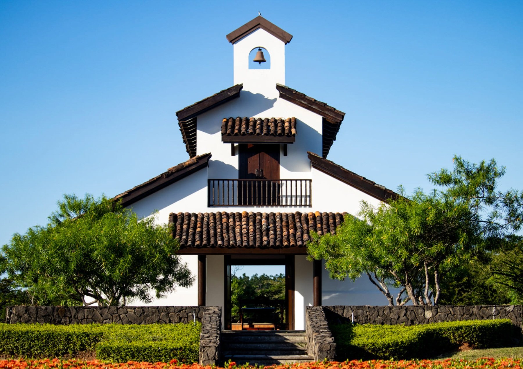 Front view of a white stucco building with a steeple, brown tiled roof, and wooden accents, surrounded by green trees and a stone wall, under a clear blue sky.