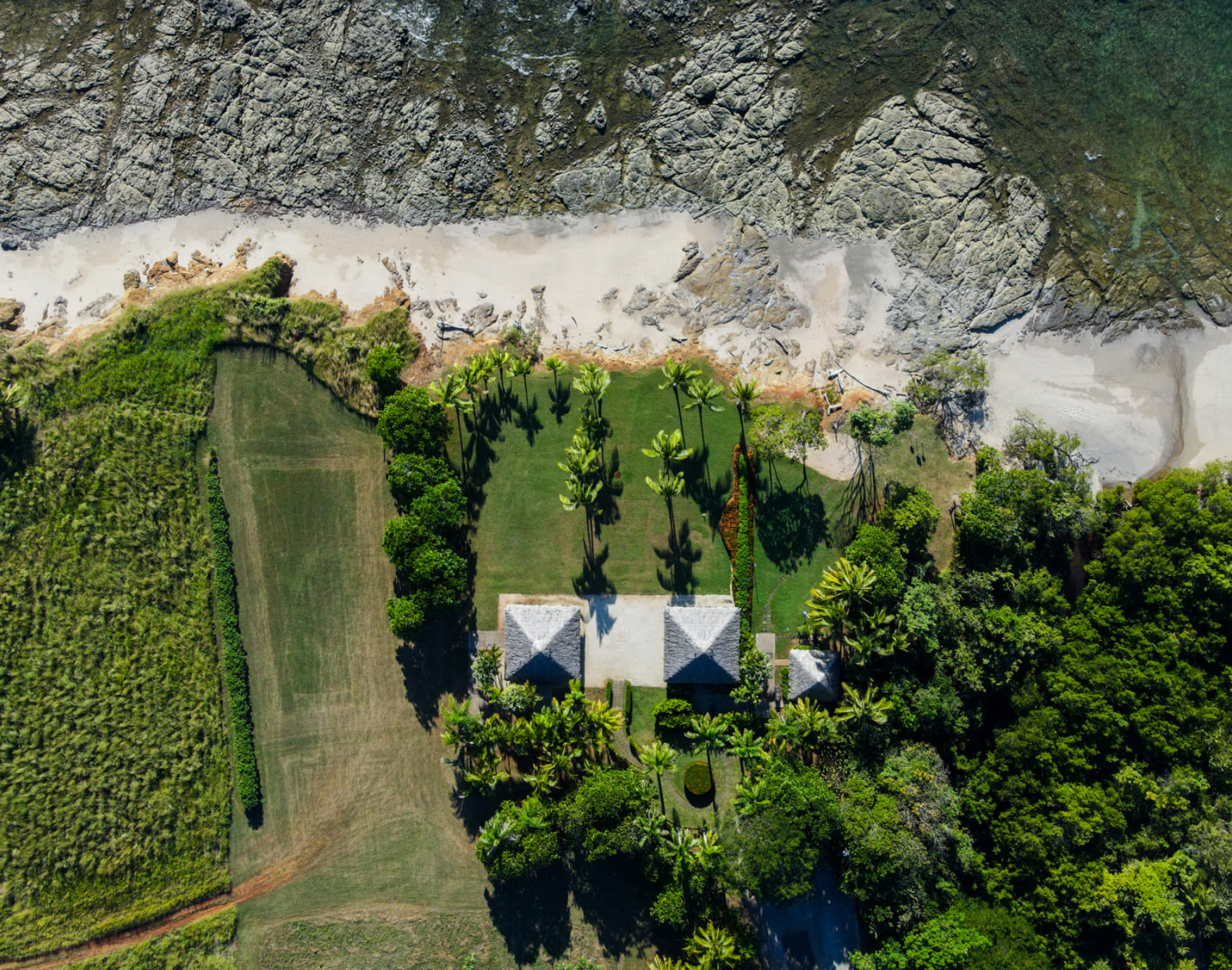 Aerial view of a tropical landscape featuring a sandy river or beach, lush green trees and vegetation, and two small white-roofed structures.