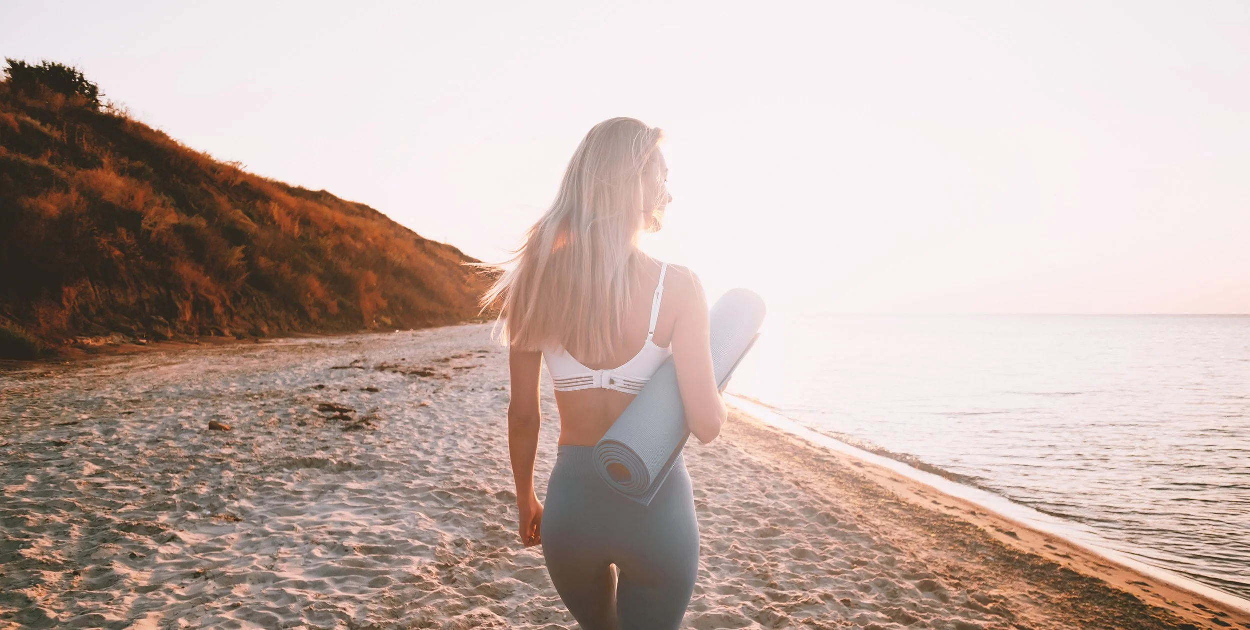 A woman with long blonde hair holding a yoga mat is walking on a sandy beach at sunset.