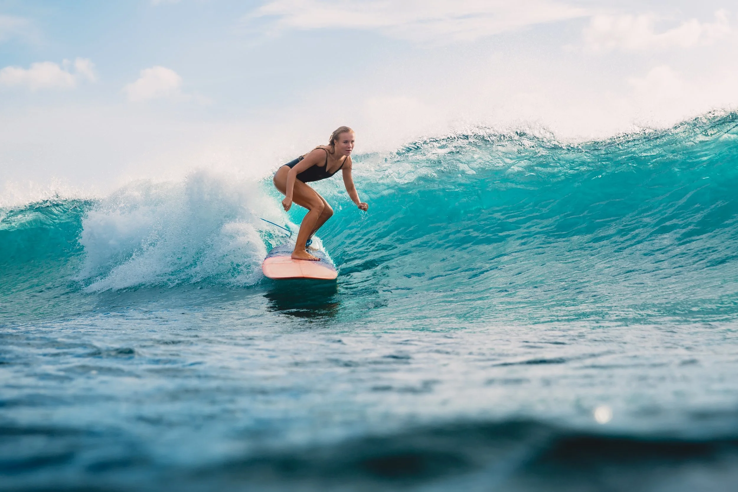 A woman surfing on a turquoise wave in the ocean, wearing a black swimsuit, with the sky and clouds in the background.