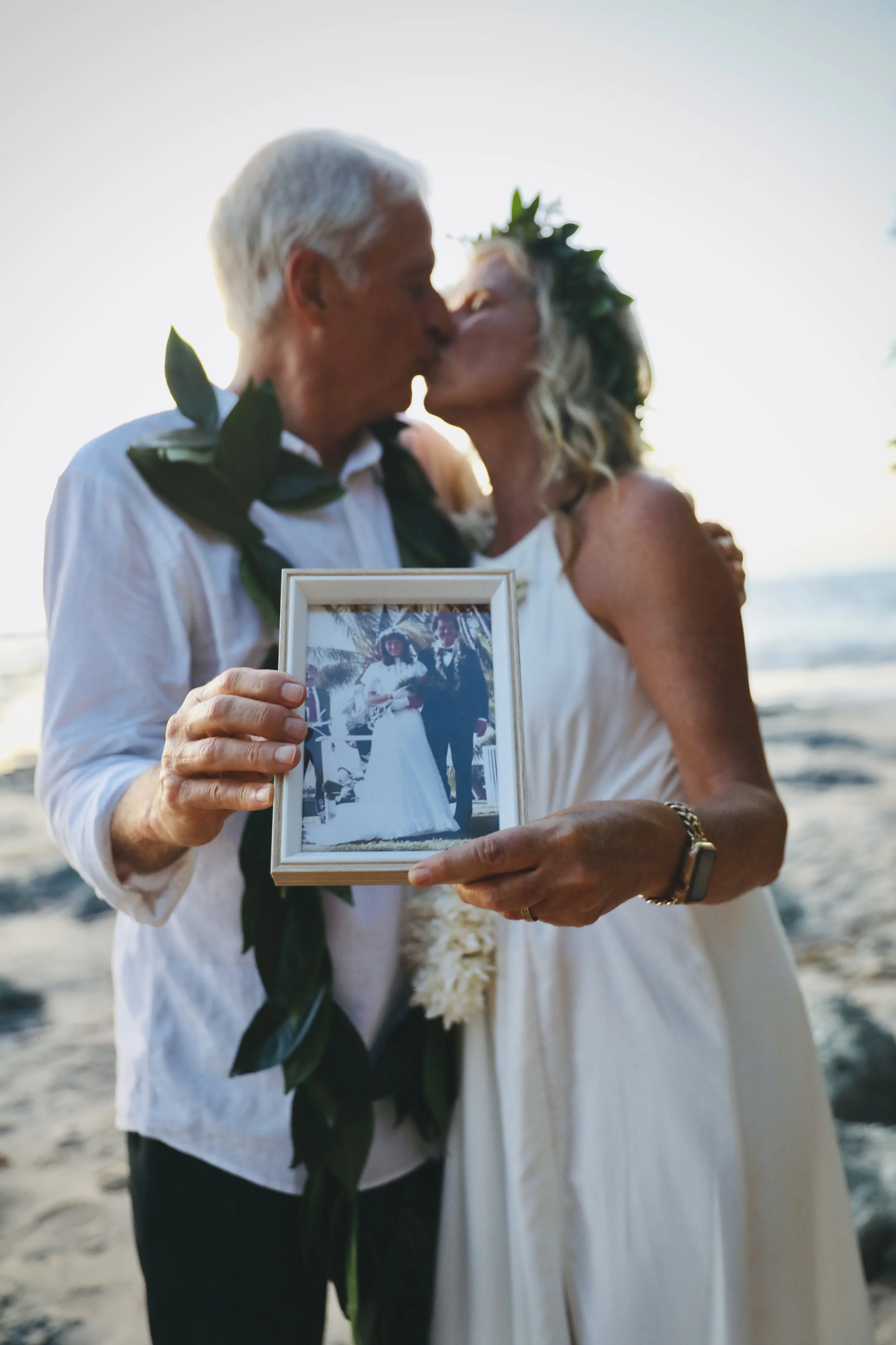 A couple, celebrating thier 40th wedding anniversary, kissing on the beach while holding a framed photograph of themselves at thier wedding.