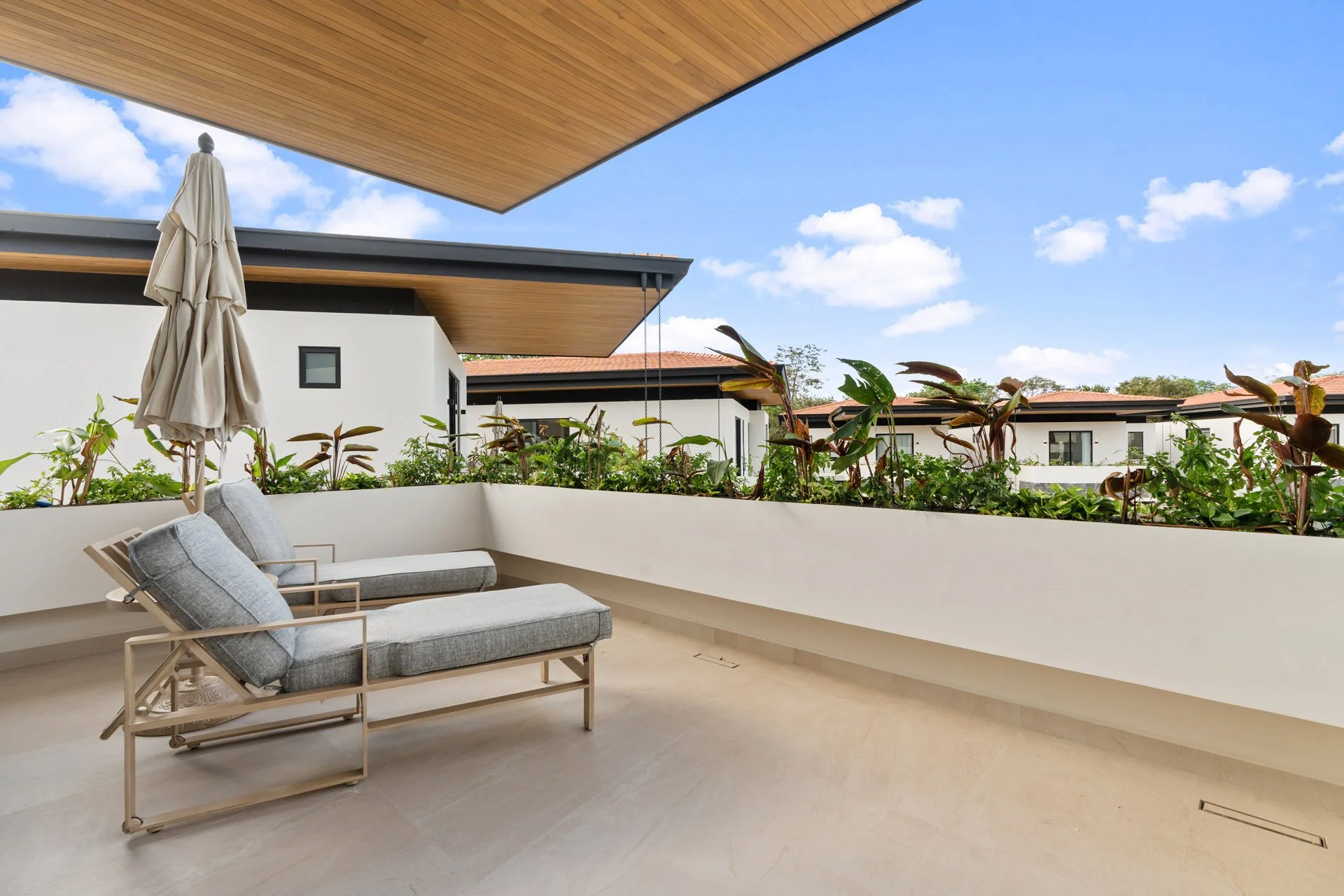 Modern outdoor balcony with two cushioned lounge chairs, a closed umbrella, and a white planter box with green plants, overlooking neighboring houses and a partly cloudy blue sky.
