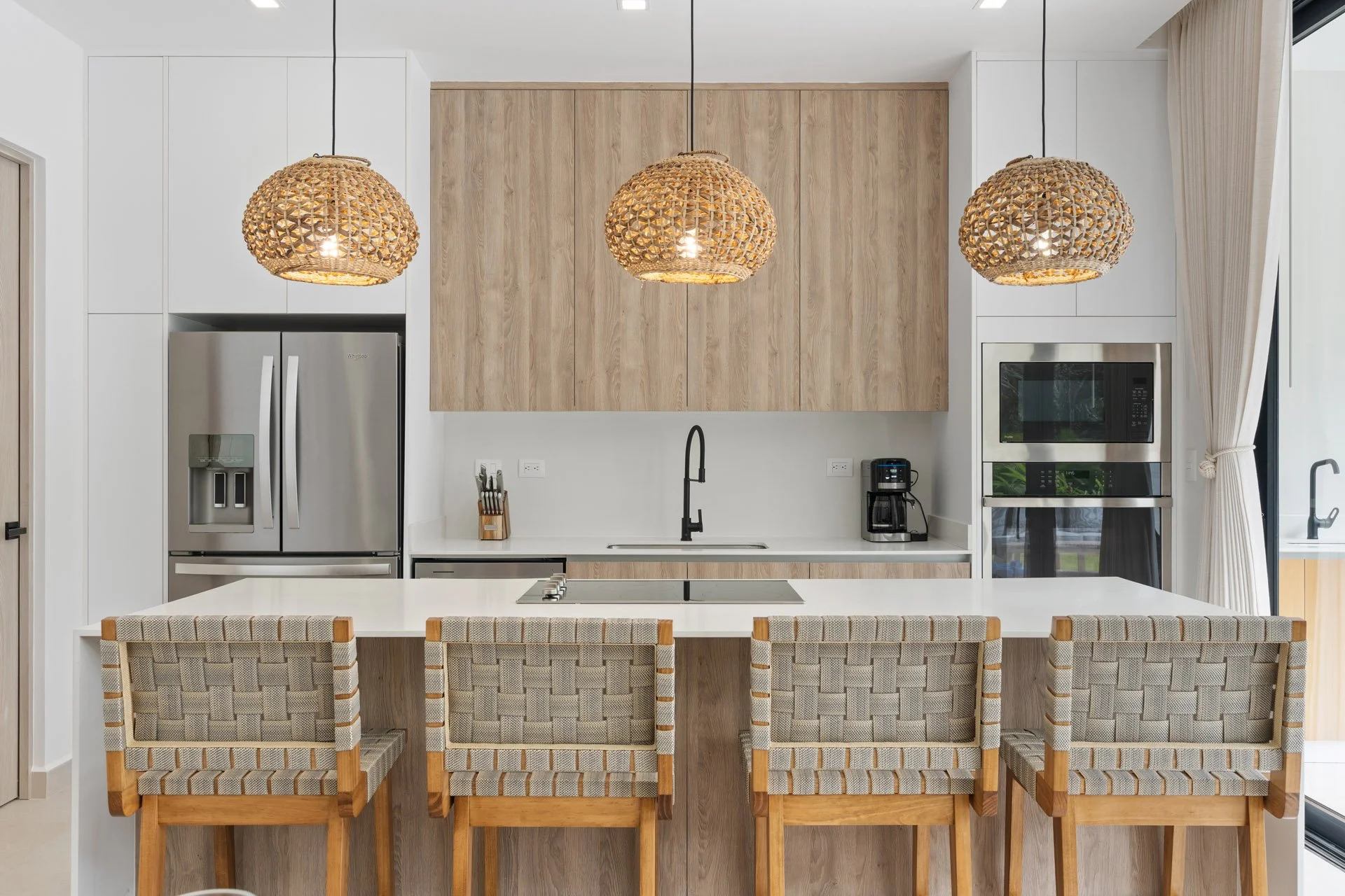 Modern kitchen with white and wood cabinets, stainless steel refrigerator, built-in microwave and oven, black faucet, coffee maker, and four woven bar stools at a white island. Three rattan pendant lights hang above.