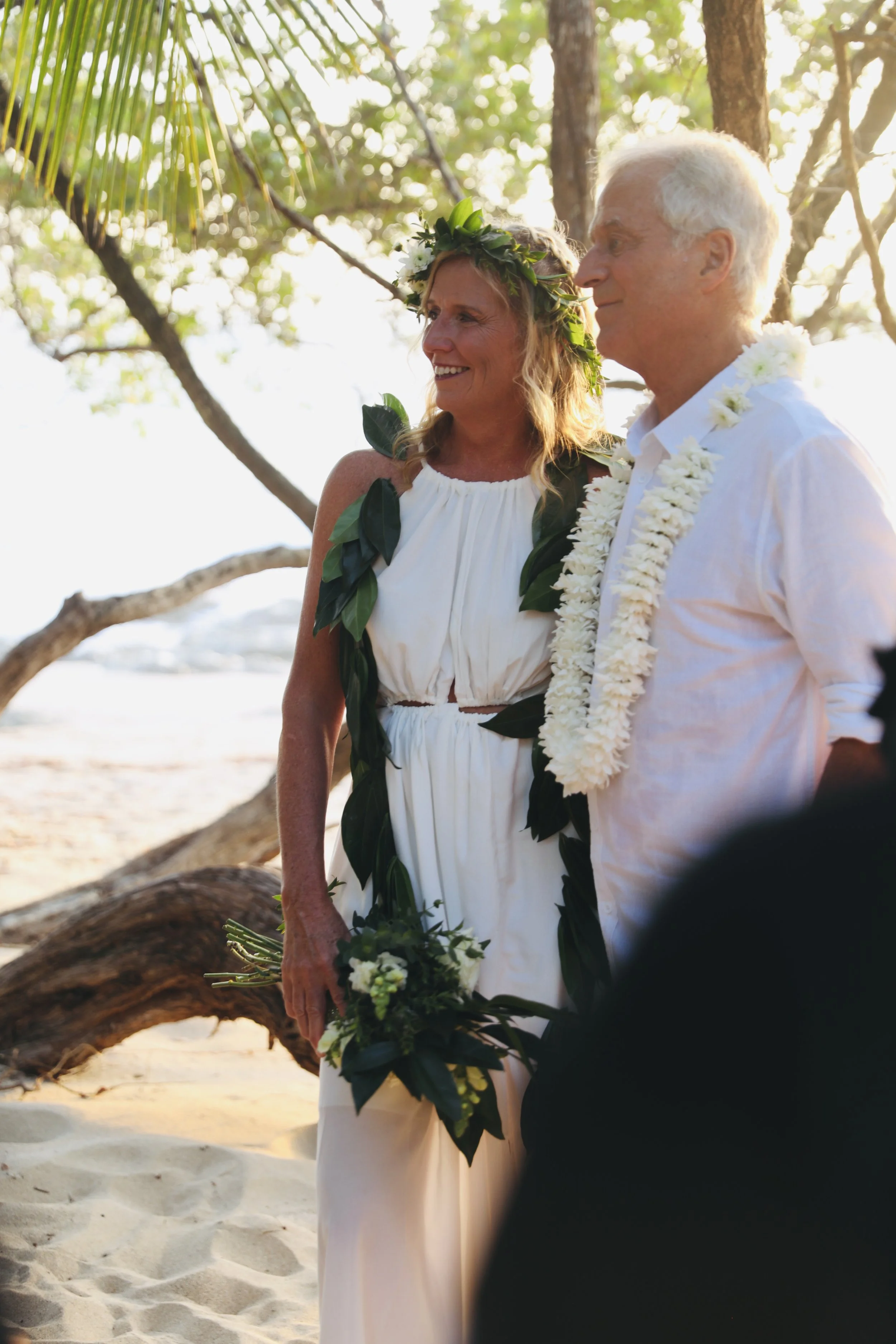 A couple gets married on a beach, dressed in white with floral leis and a greenery crown for the woman. They stand near trees and driftwood, smiling during sunset.