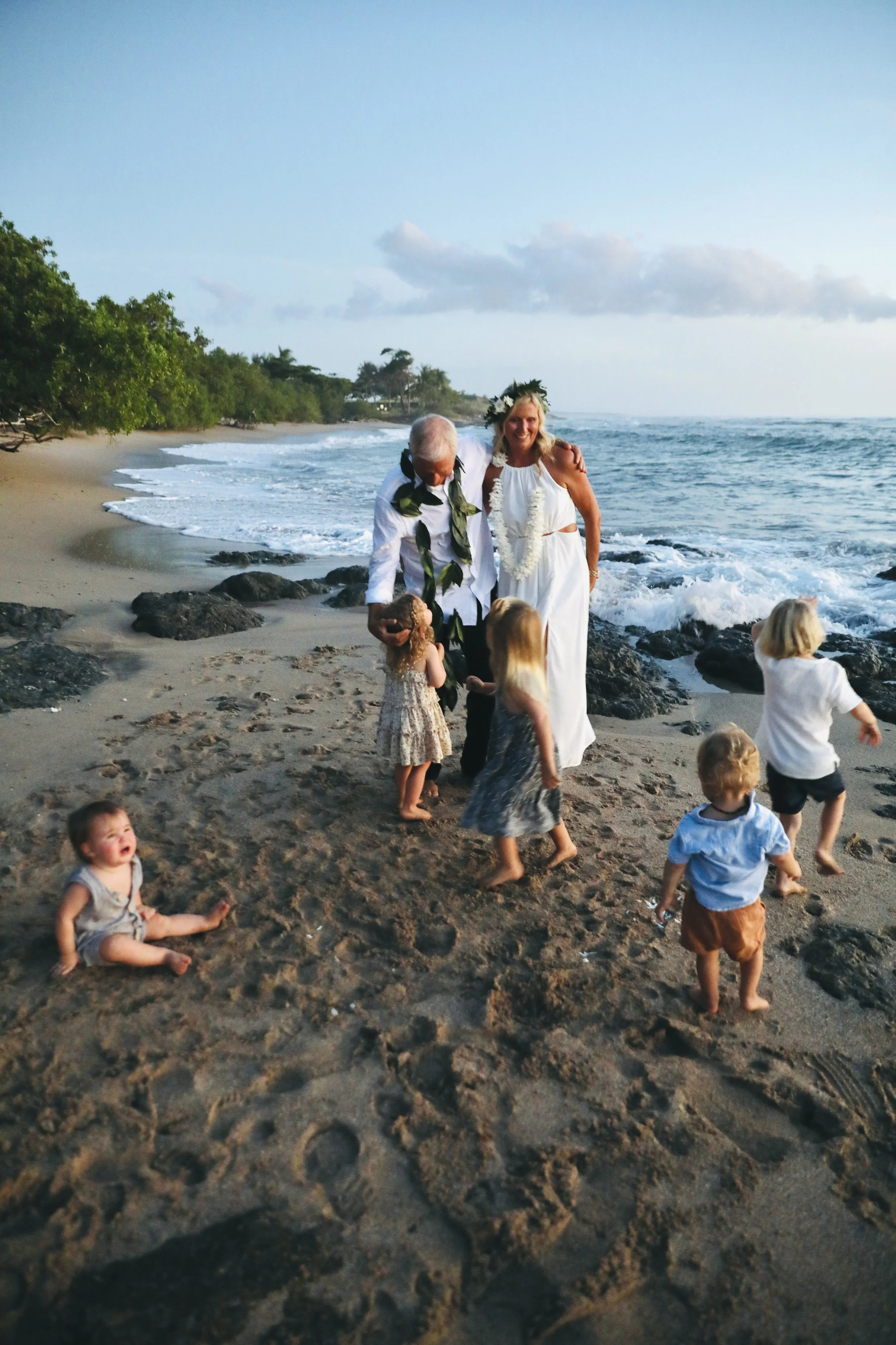 A beach wedding scene with a couple in white attire, surrounded by small children on the sand near the ocean at sunset.