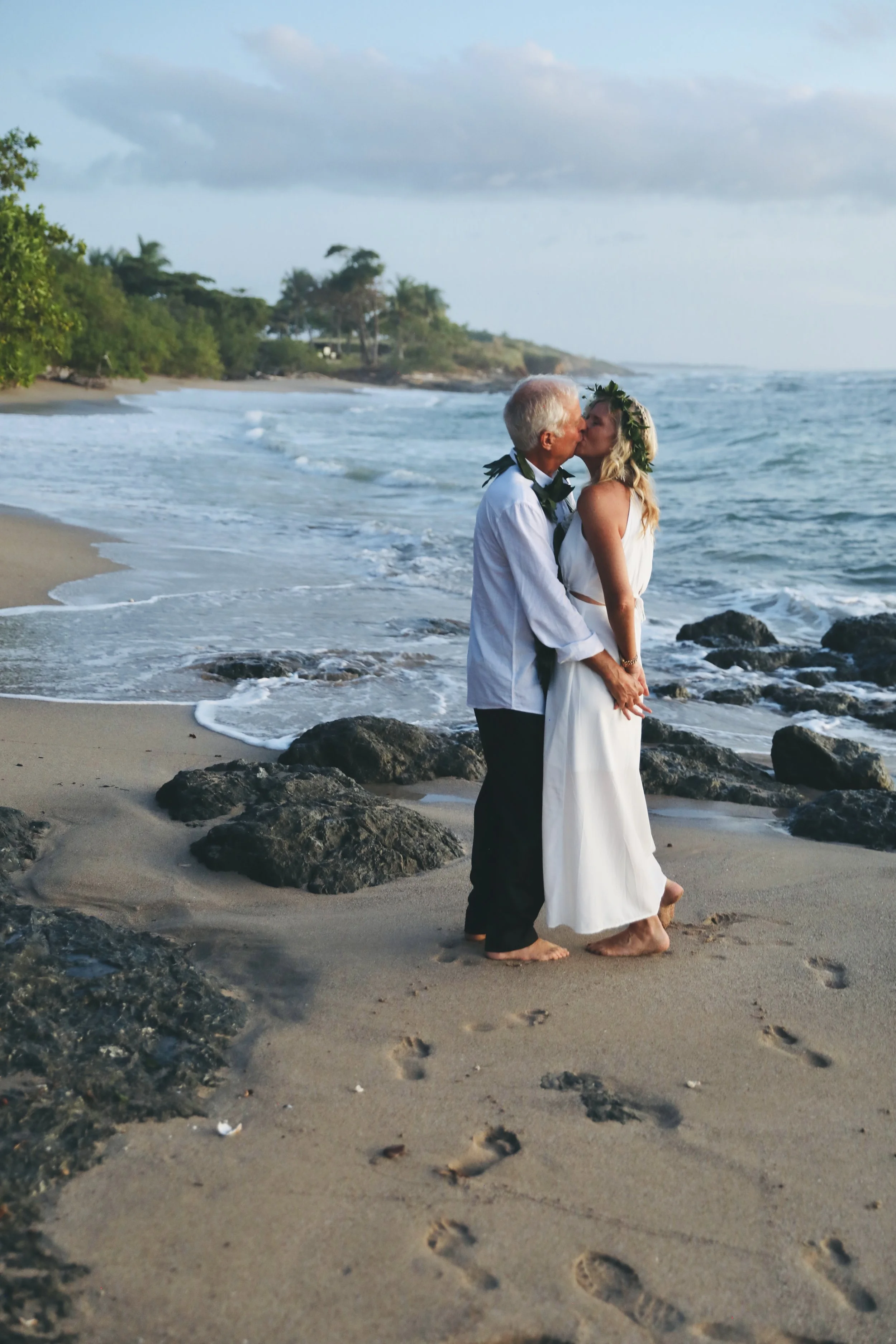 A happy couple are kissing on a sandy beach, dressed in wedding attire, with the ocean and rocks in the background.