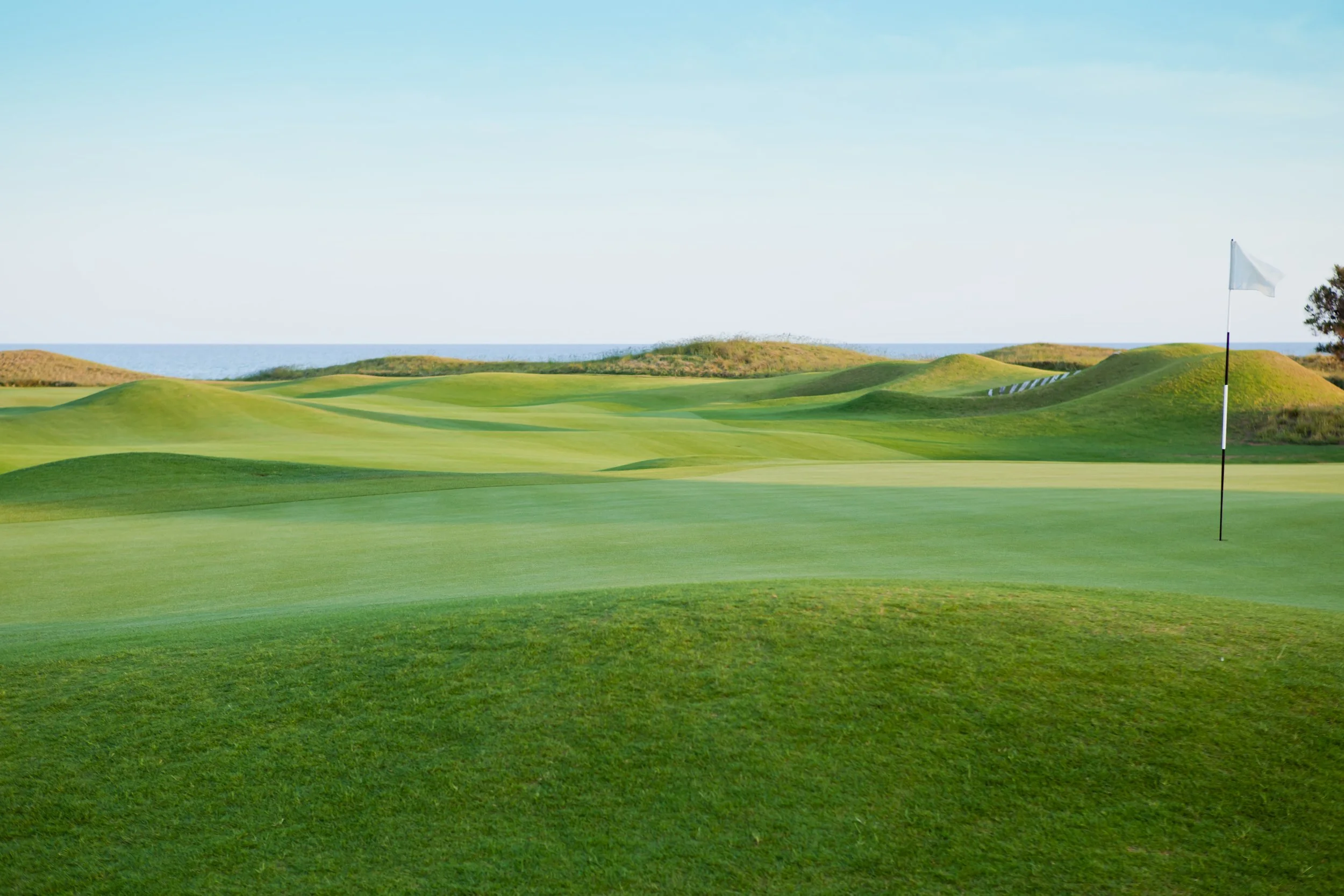 A lush golf course with green fairways, rolling hills, and a white flag on a green hole under a clear blue sky.