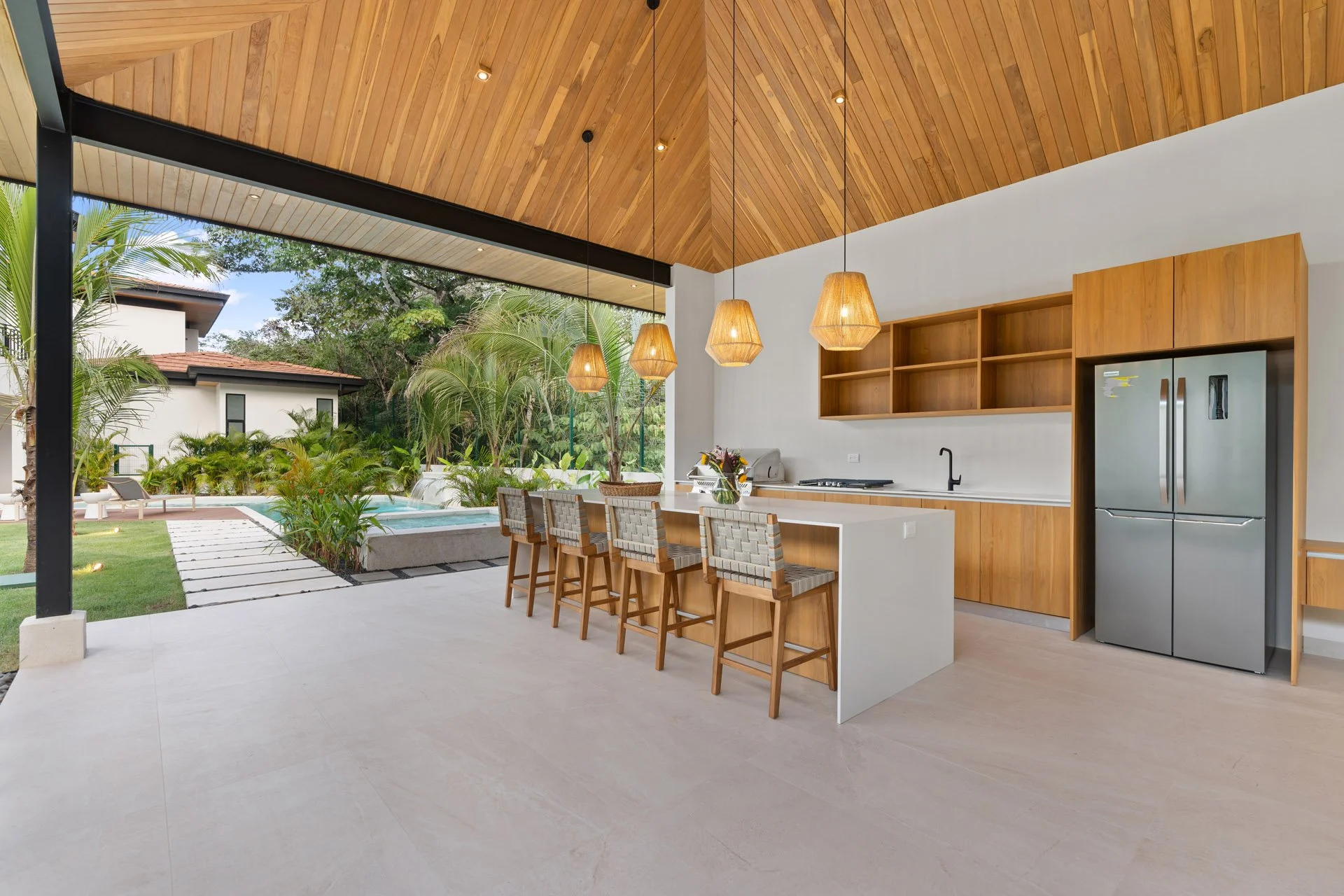 Modern open-concept kitchen with wooden cabinetry, a large kitchen island with bar seating, pendant lighting, and a view of a backyard pool area with trees and lounge chairs.