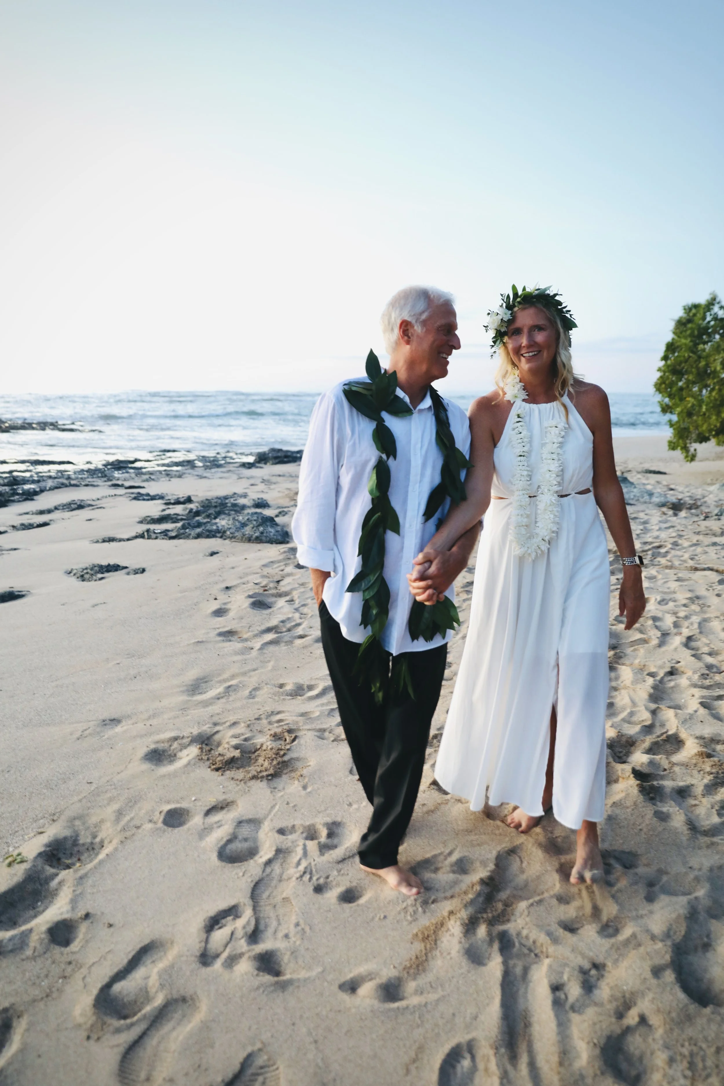 A couple dressed in white wearing leis and flower crowns, holding hands and walking barefoot on the beach during sunset.
