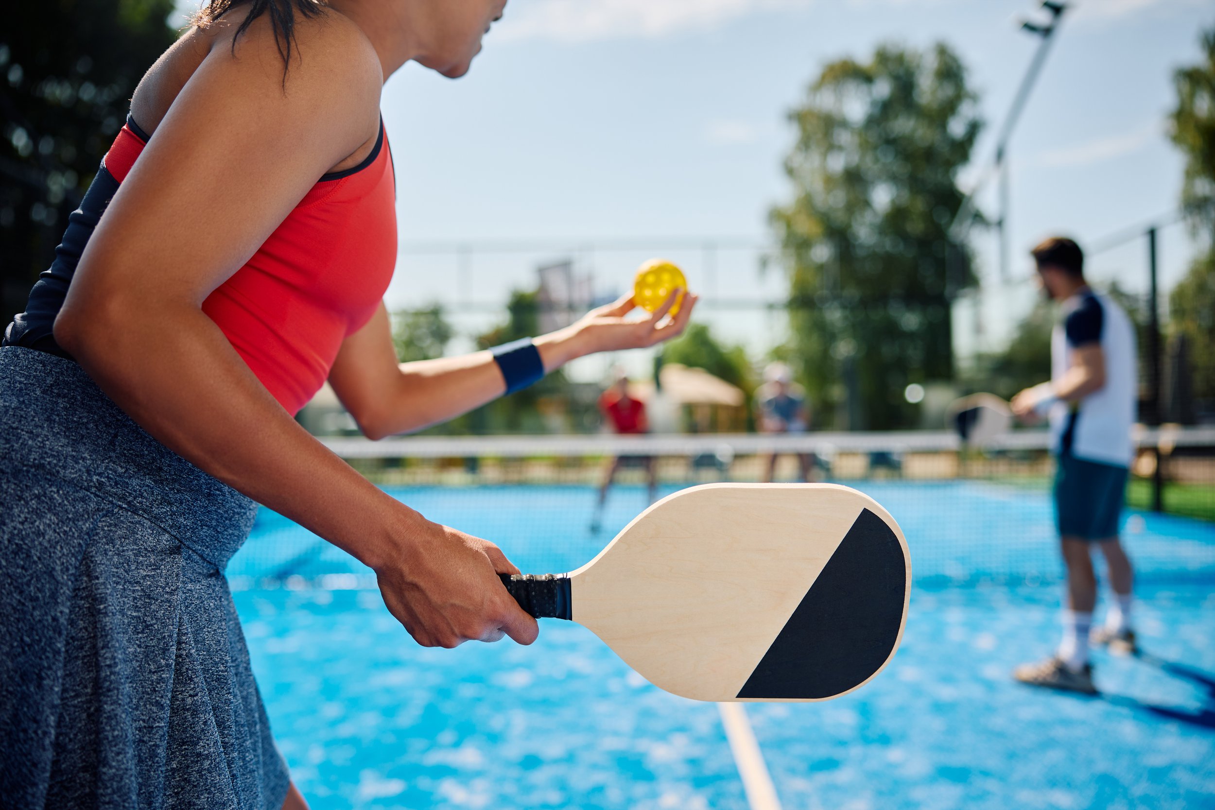 A woman playing pickleball on an outdoor court, holding a paddle and a yellow pickleball, with other players in the background.