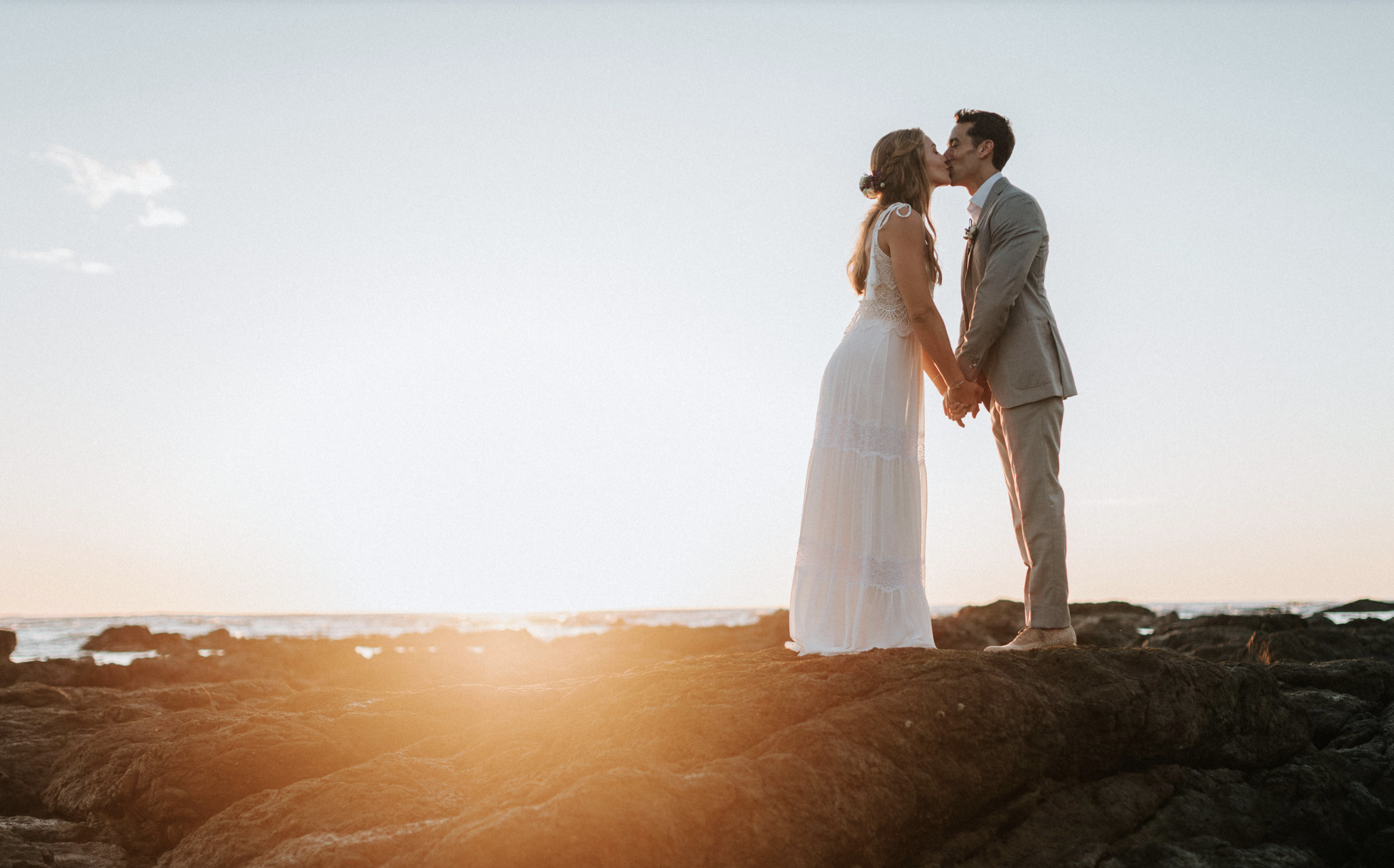 A bride and groom sharing a kiss and holding hands on rocky shoreline at sunset.