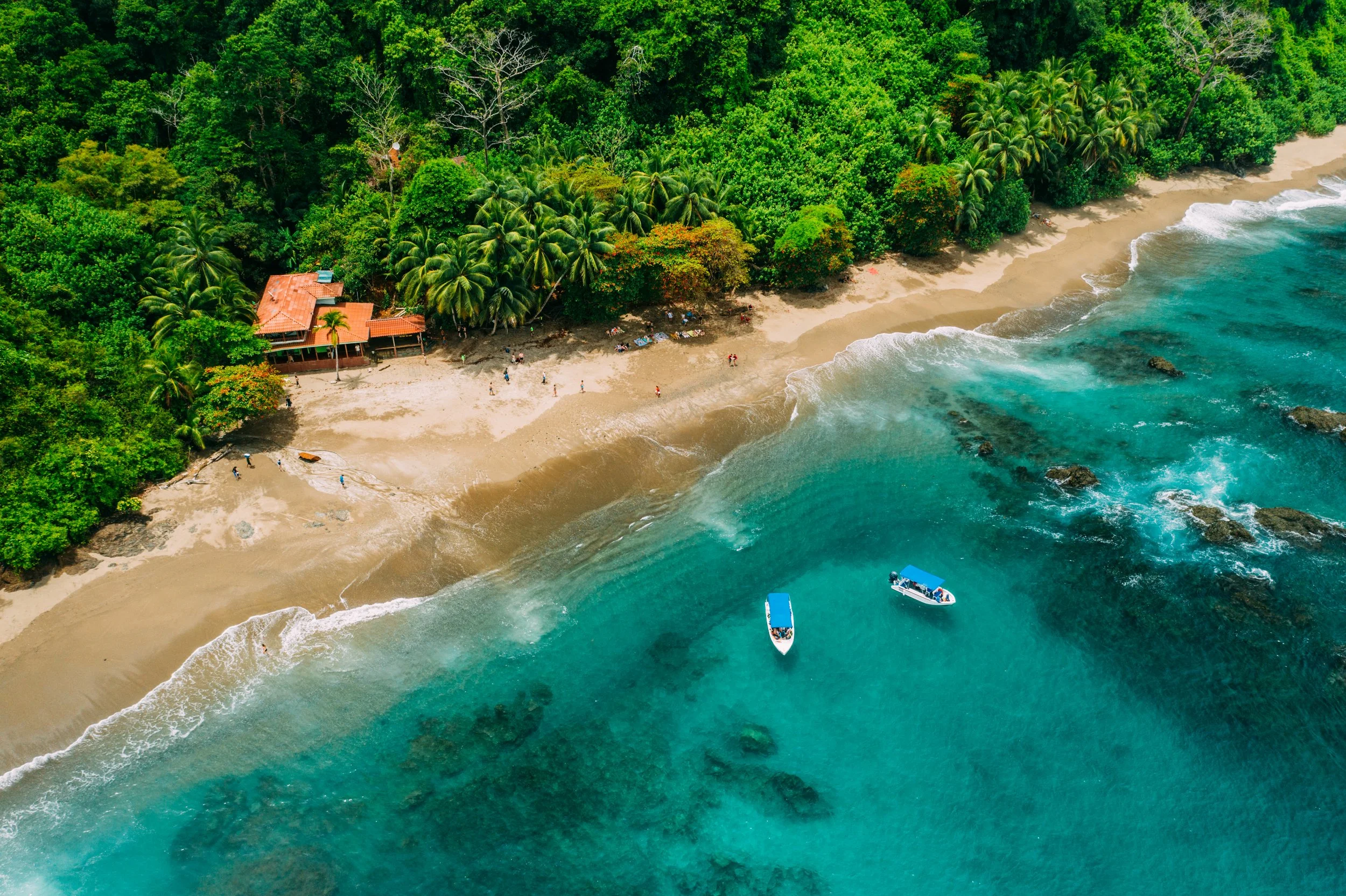 Aerial view of a tropical beach with a dense green forest, sandy shoreline, and turquoise ocean waters with two boats.