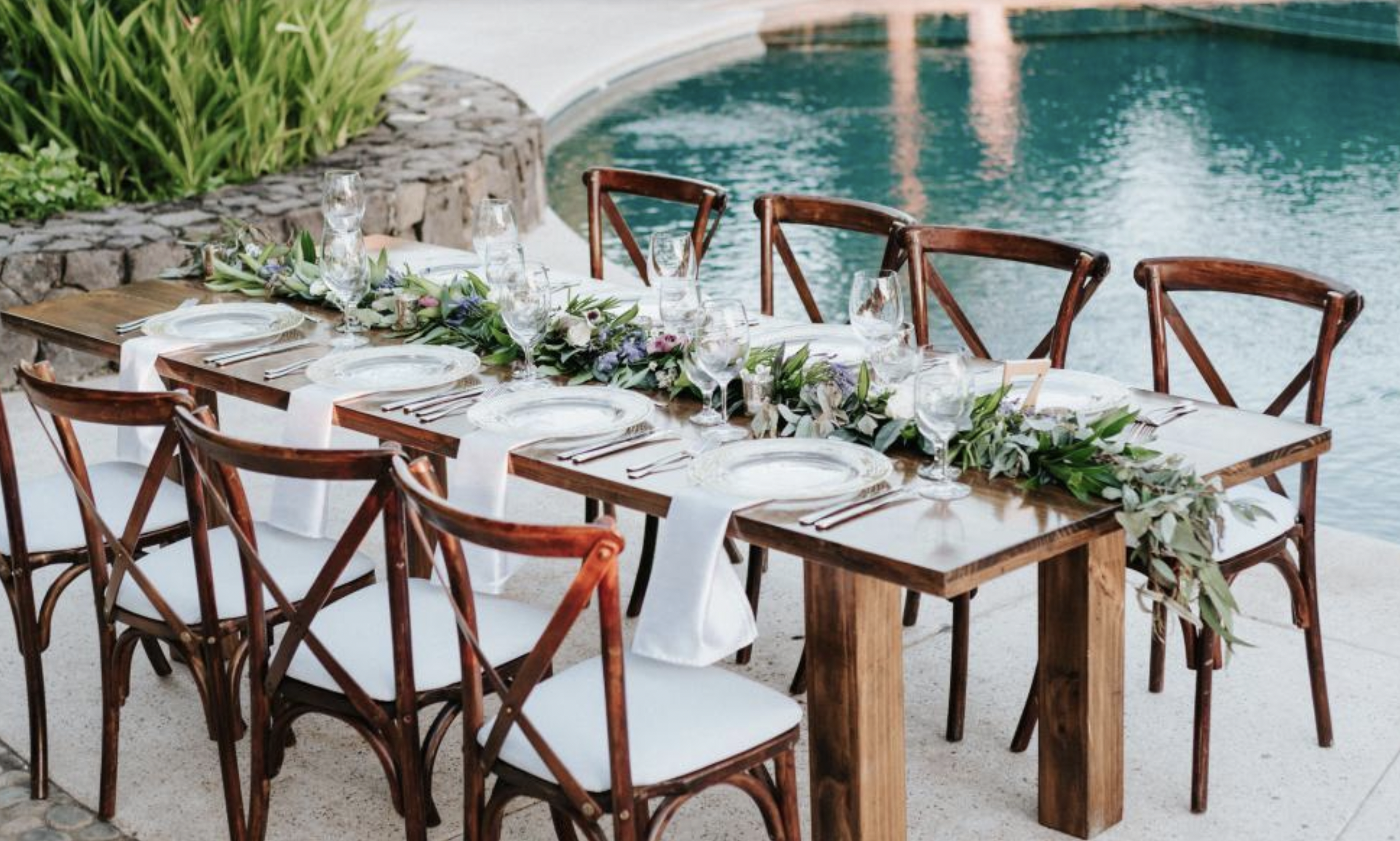 A long wooden dining table set for a formal outdoor meal near a swimming pool, with white tablecloths, greenery, and floral centerpieces, surrounded by wooden chairs.