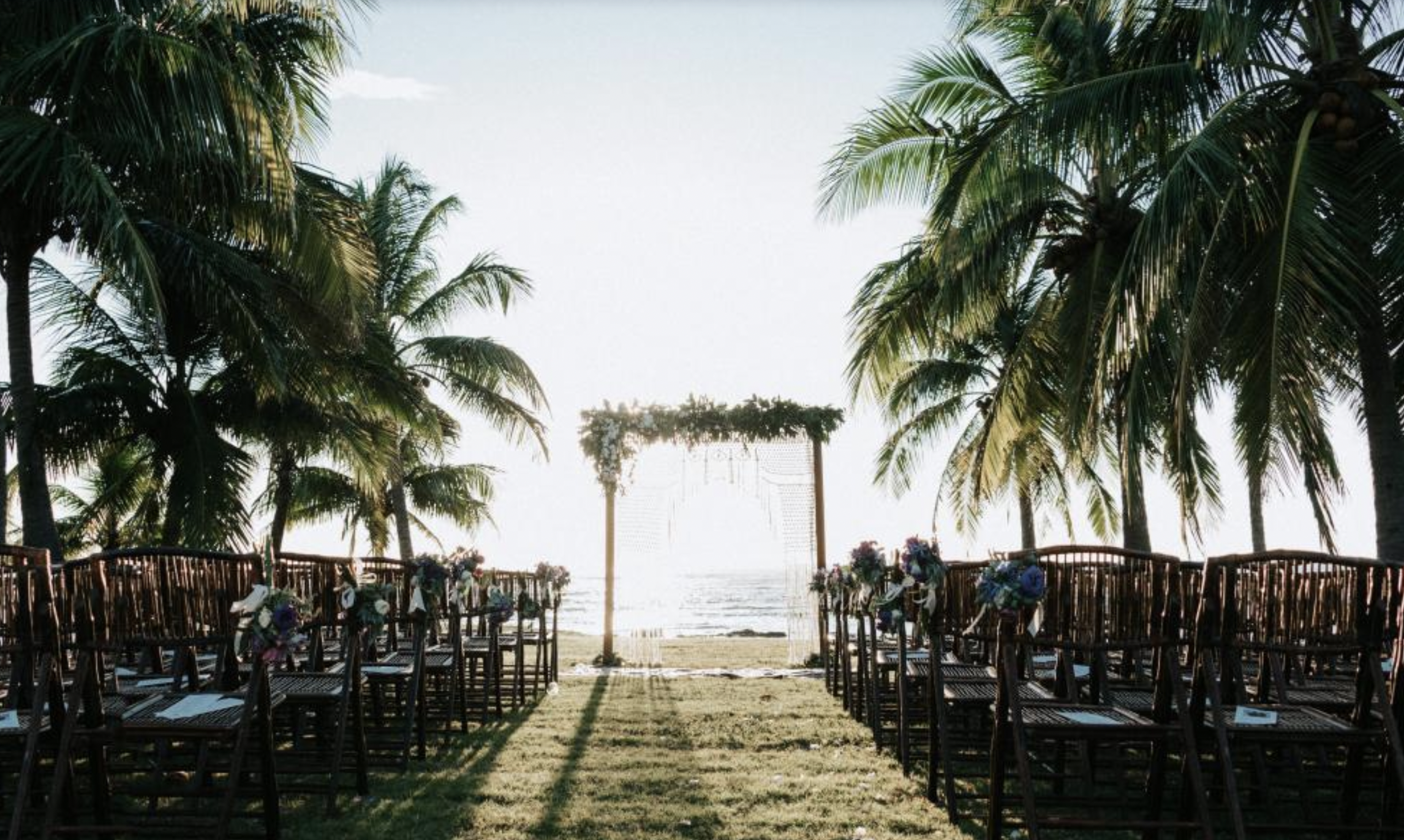 Beachside wedding setup with wooden chairs decorated with flowers, facing a wooden arch draped with greenery, under palm trees by the ocean.