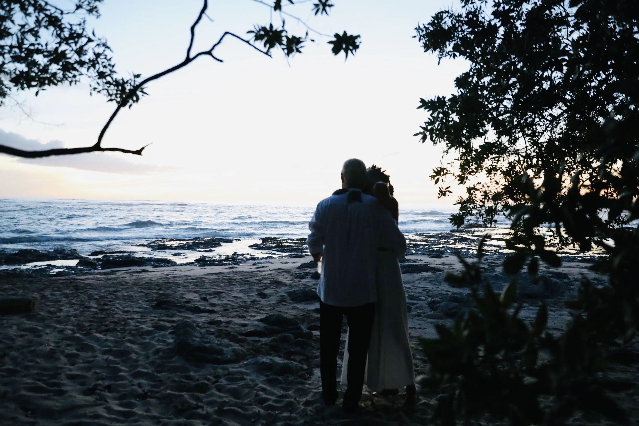 A couple embracing on a beach at sunset, surrounded by trees and rocks in the foreground.