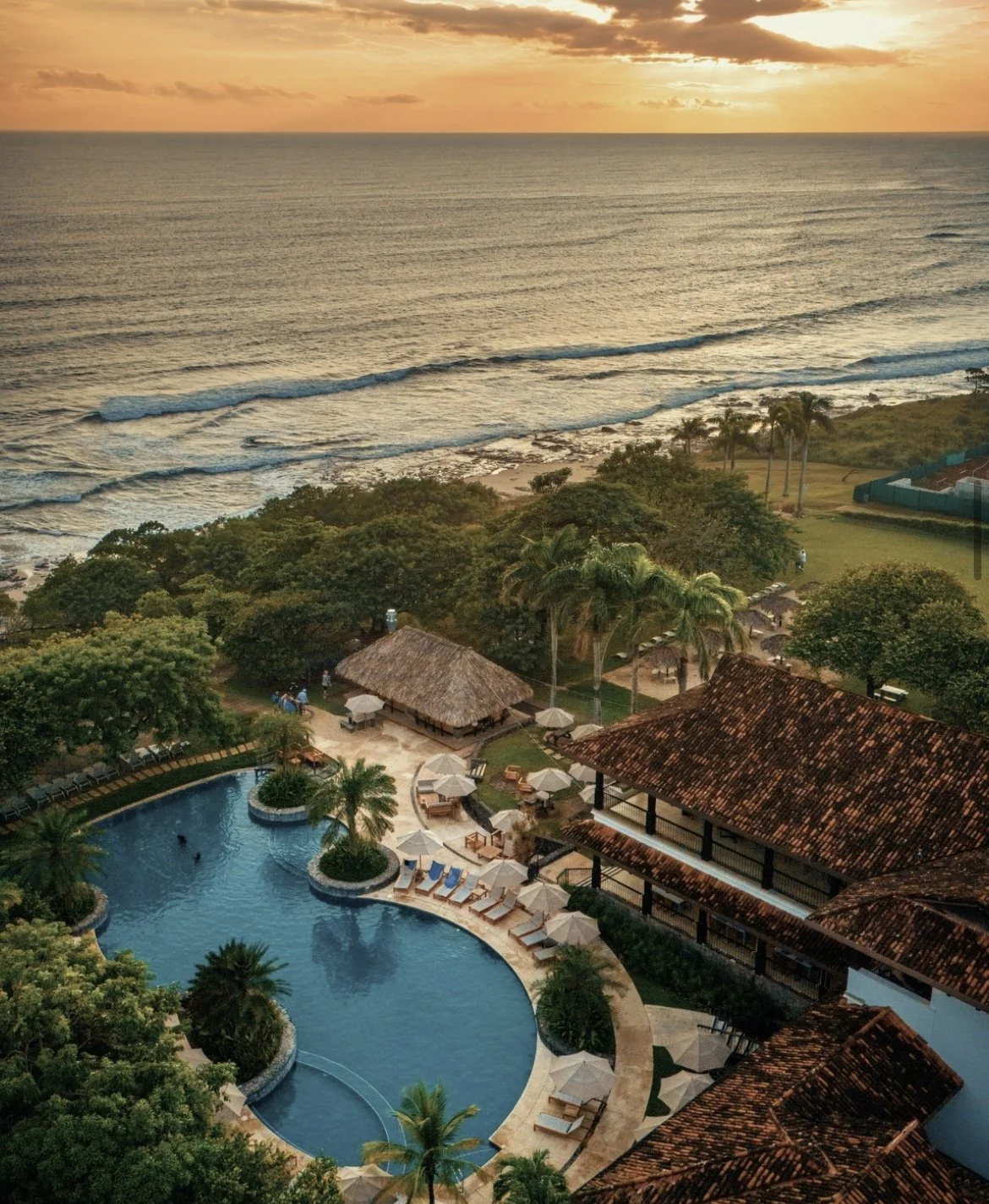 Aerial view of a tropical resort at sunset featuring a swimming pool, palm trees, thatched-roof structures, and a beach with ocean waves in the background.