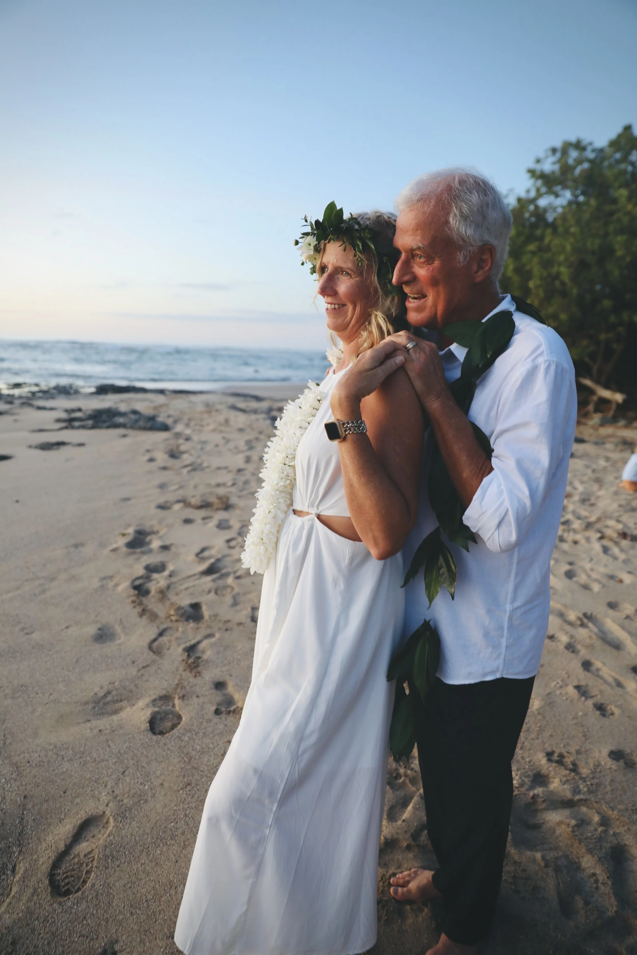 A couple in wedding attire hugging on a beach at sunset, with ocean waves and trees in the background.
