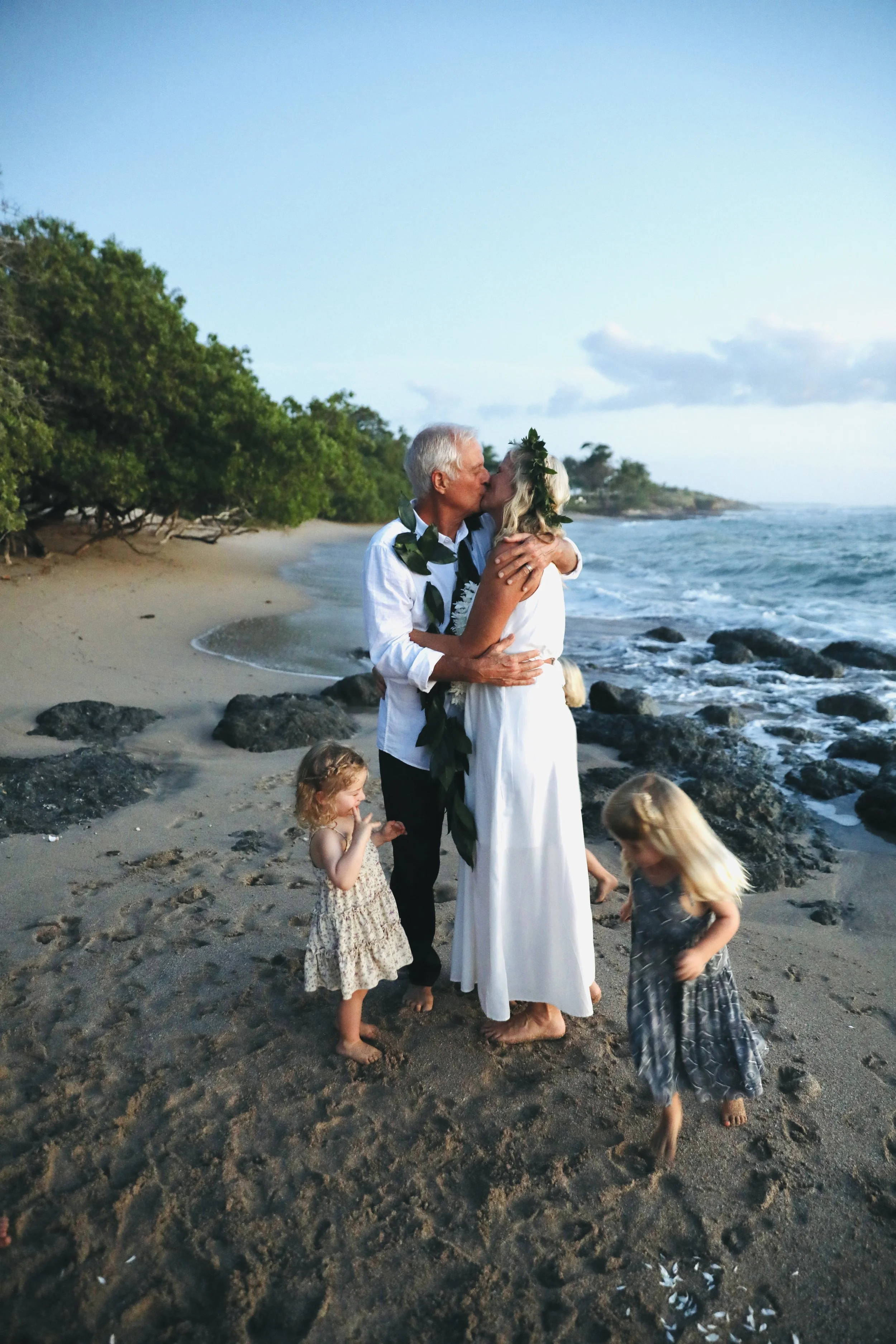 An elderly couple is kissing on a beach at sunset, surrounded by children, with waves and trees in the background.