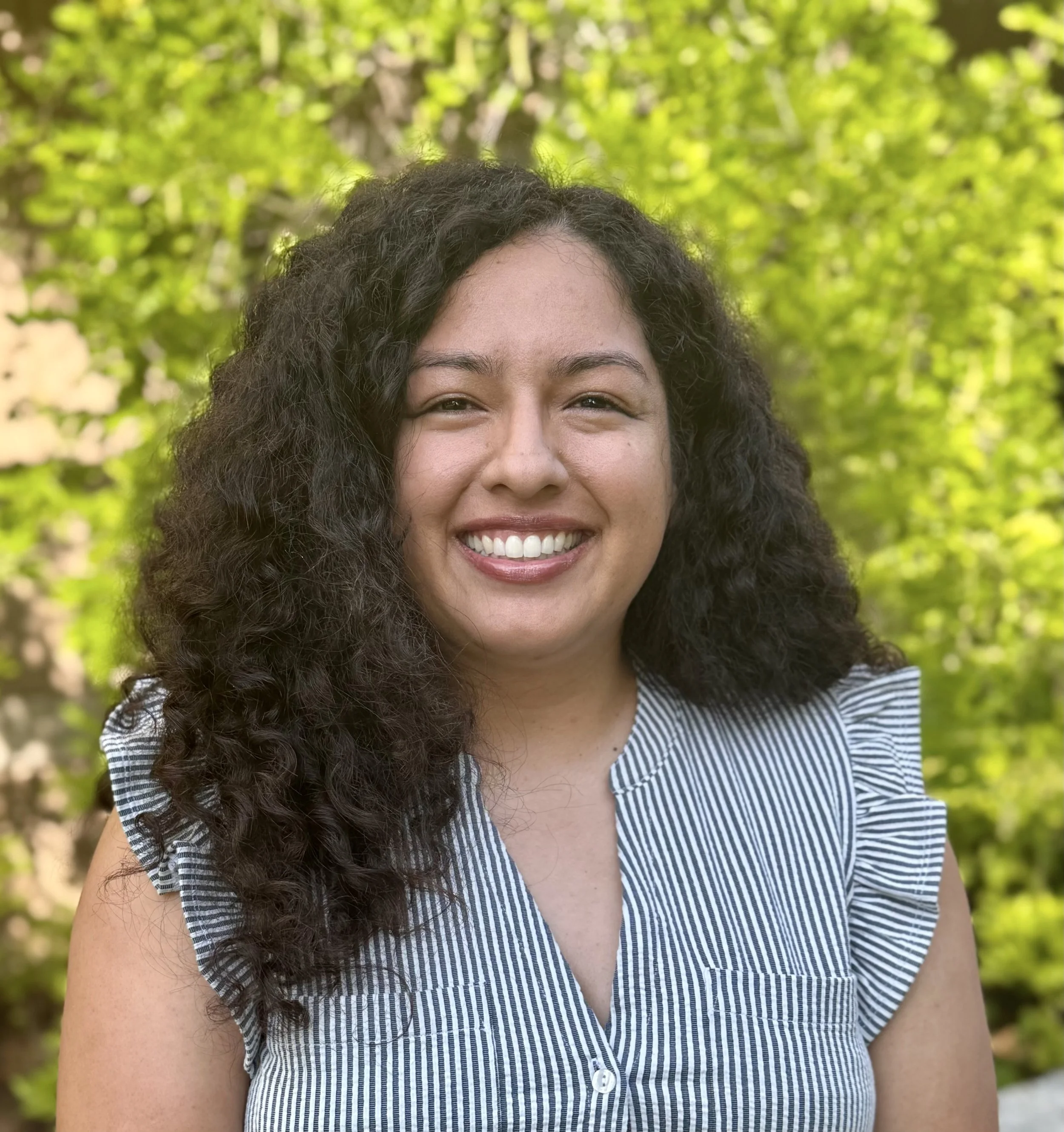 Close-up of a Latina woman smiling outdoors, curly dark hair, wearing a striped shirt, green leafy background.