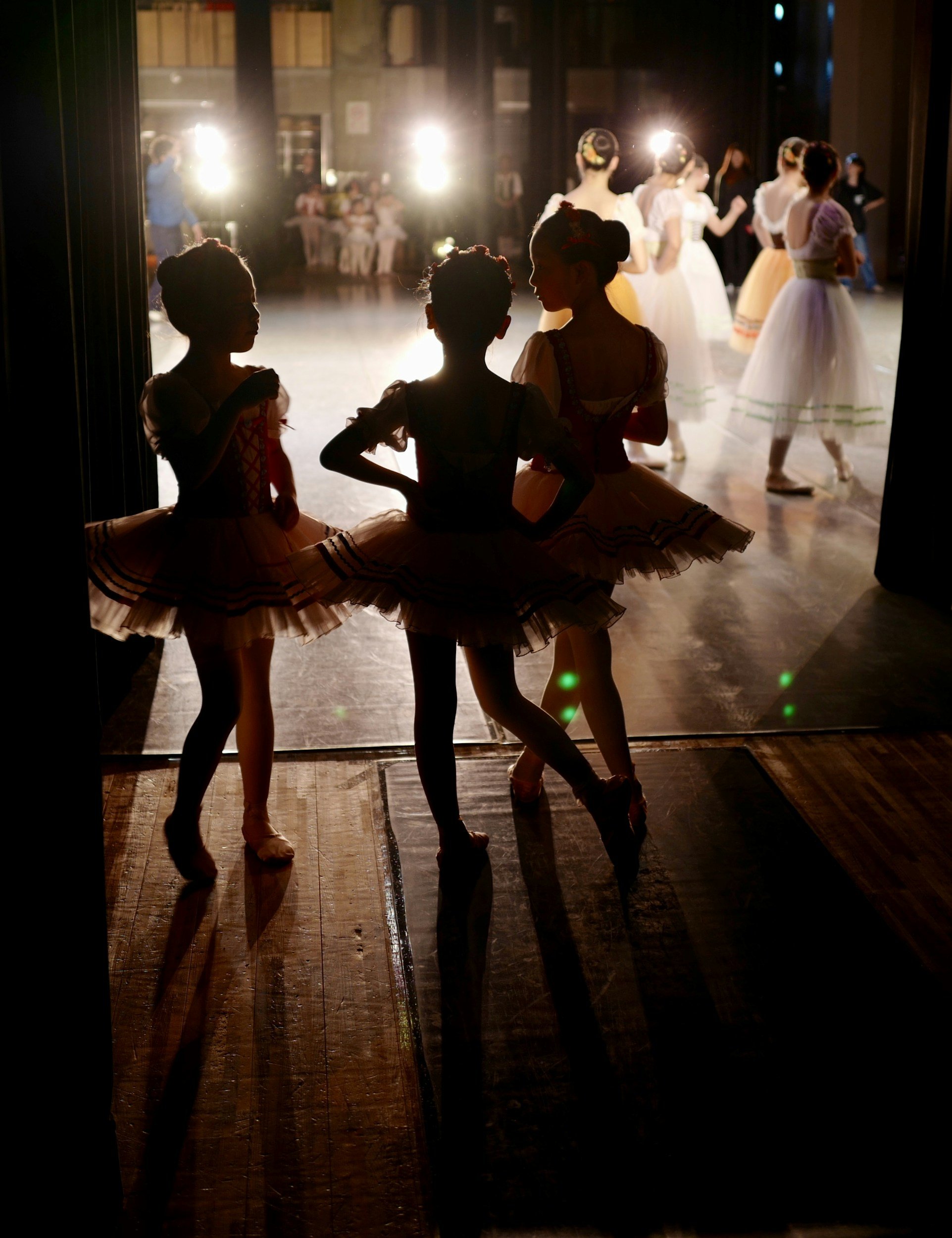 Backstage view of ballet dancers in tutus preparing to perform, with some dancers visible through an open door and stage lights illuminating the scene.