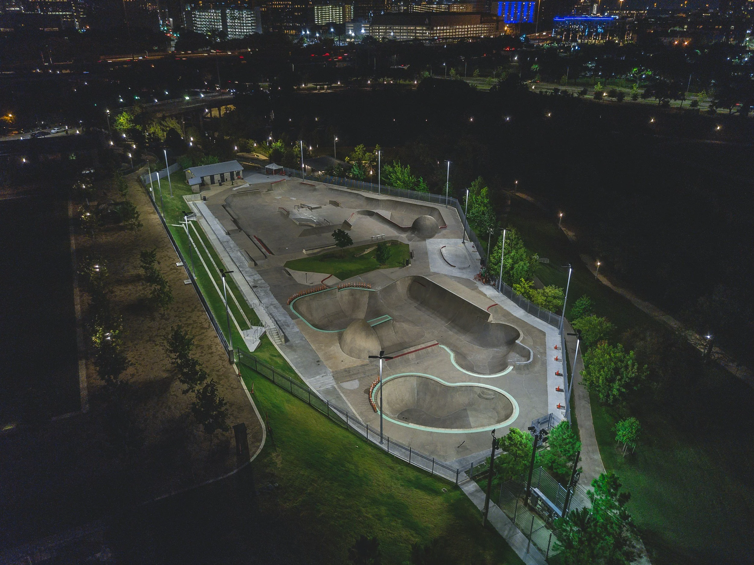 Night view of an outdoor skatepark with various ramps and bowls, illuminated by nearby streetlights, with city buildings and lights in the background.