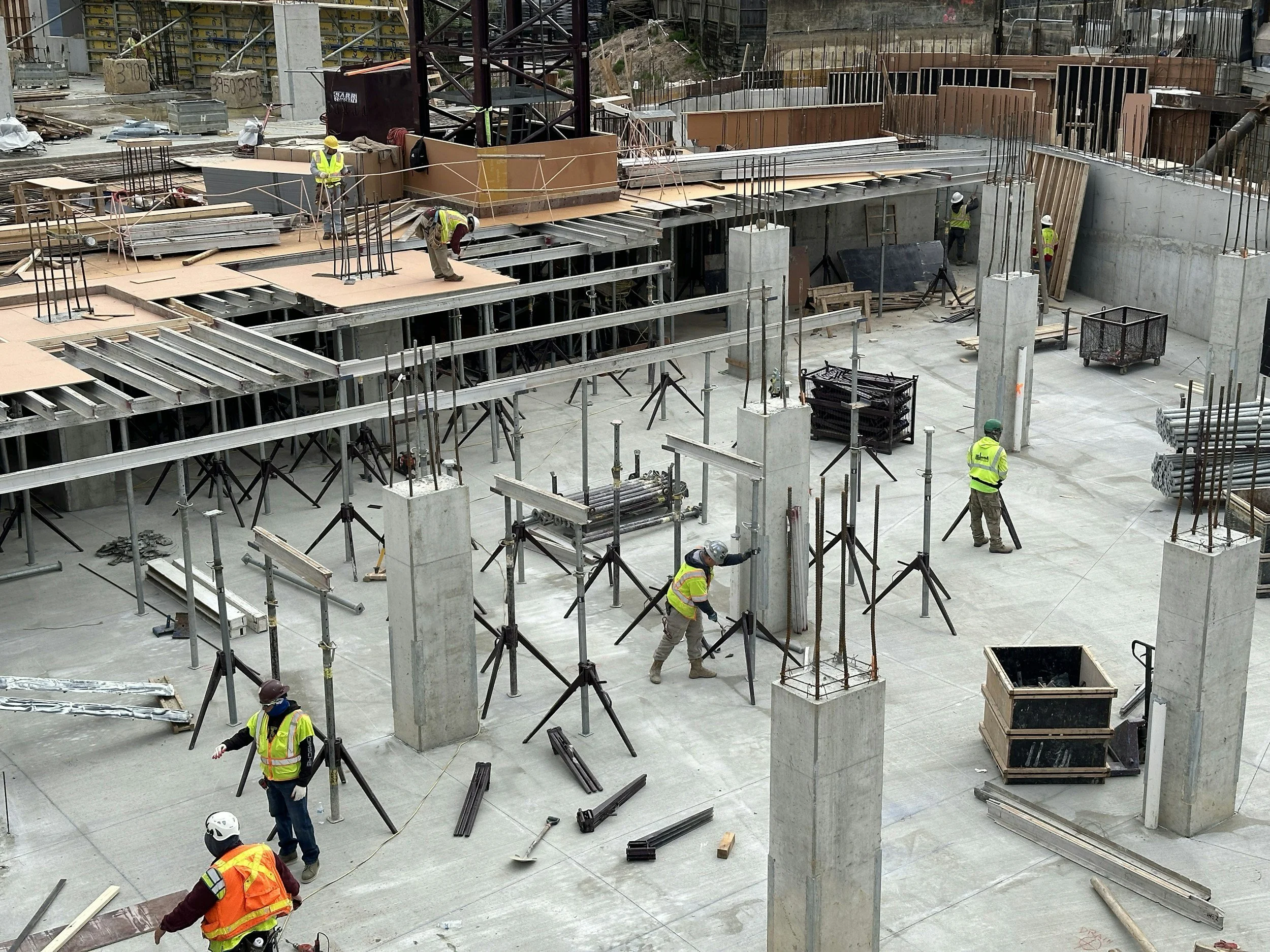Construction site with workers in yellow safety vests and hard hats working on building framework, steel and concrete structures, various construction materials and equipment scattered around.