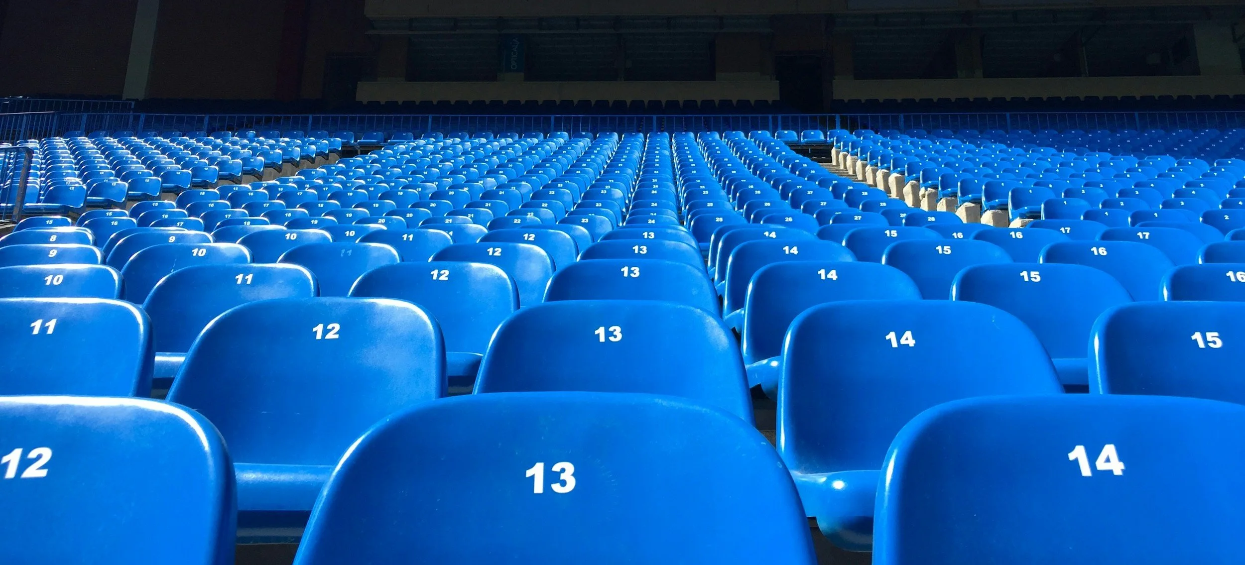 Empty blue stadium seats with numbered labels, viewed from the front looking towards the back, possibly during daytime.