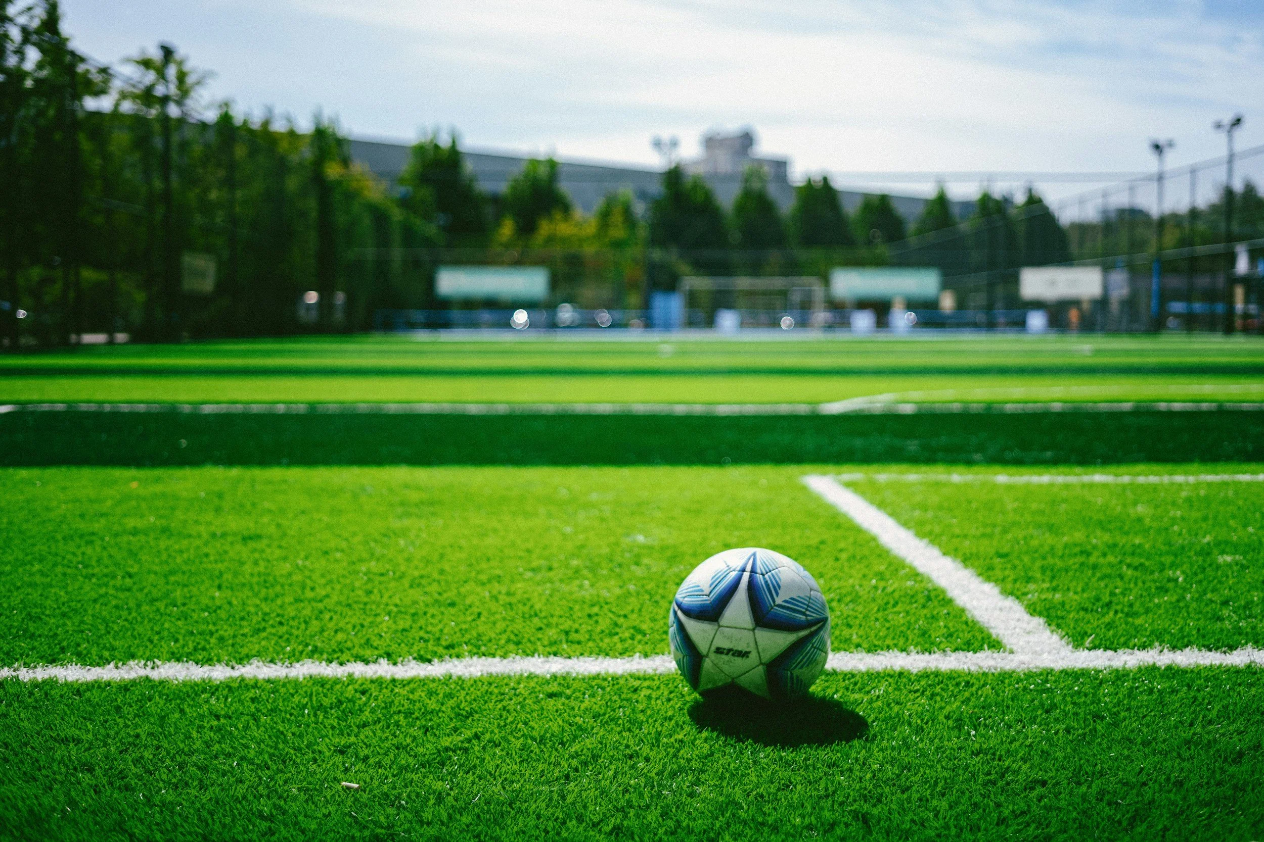 A soccer ball positioned on a green grass field with white lines, with a blurred background of trees and a sports facility under a partly cloudy sky.