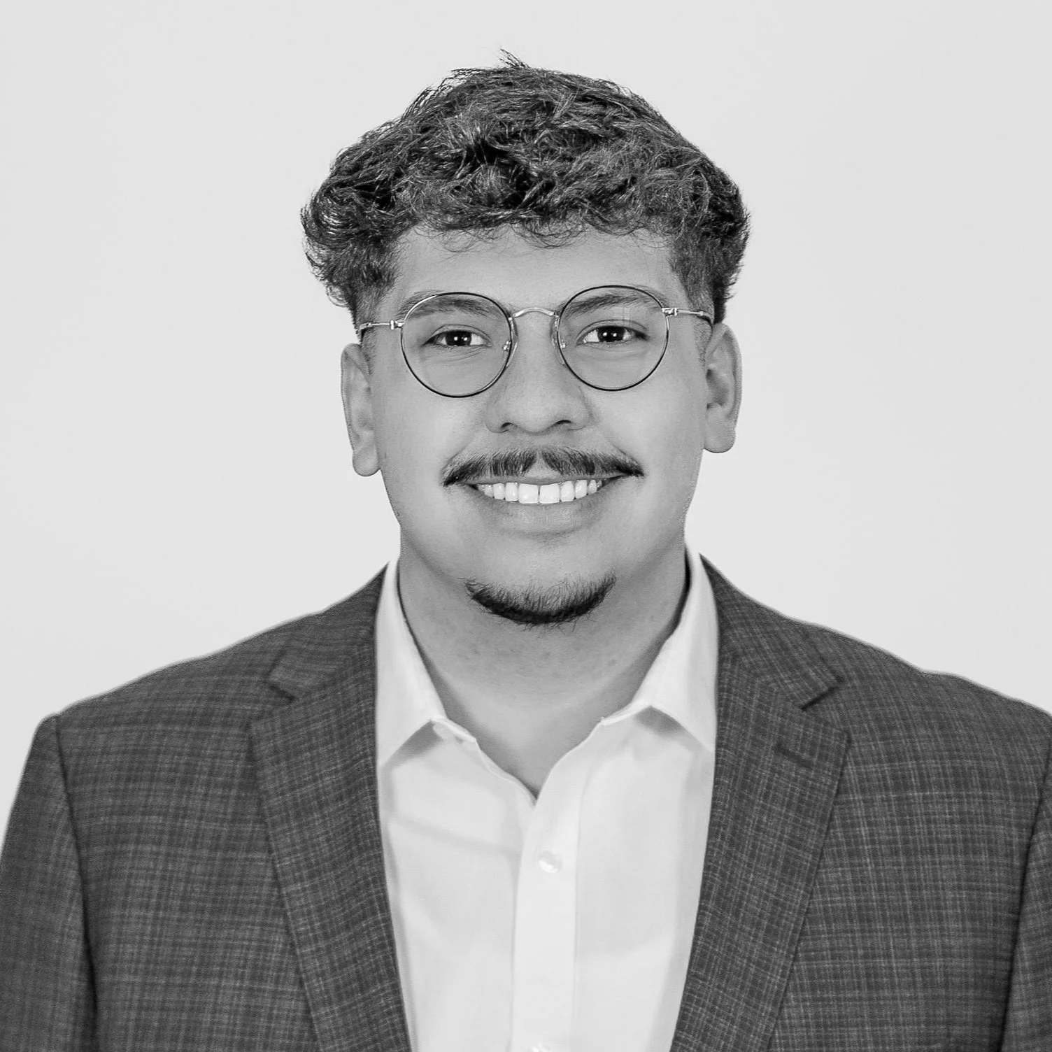 Black and white professional headshot of a young man with glasses, a mustache, and a goatee, smiling and wearing a checked blazer and white shirt.