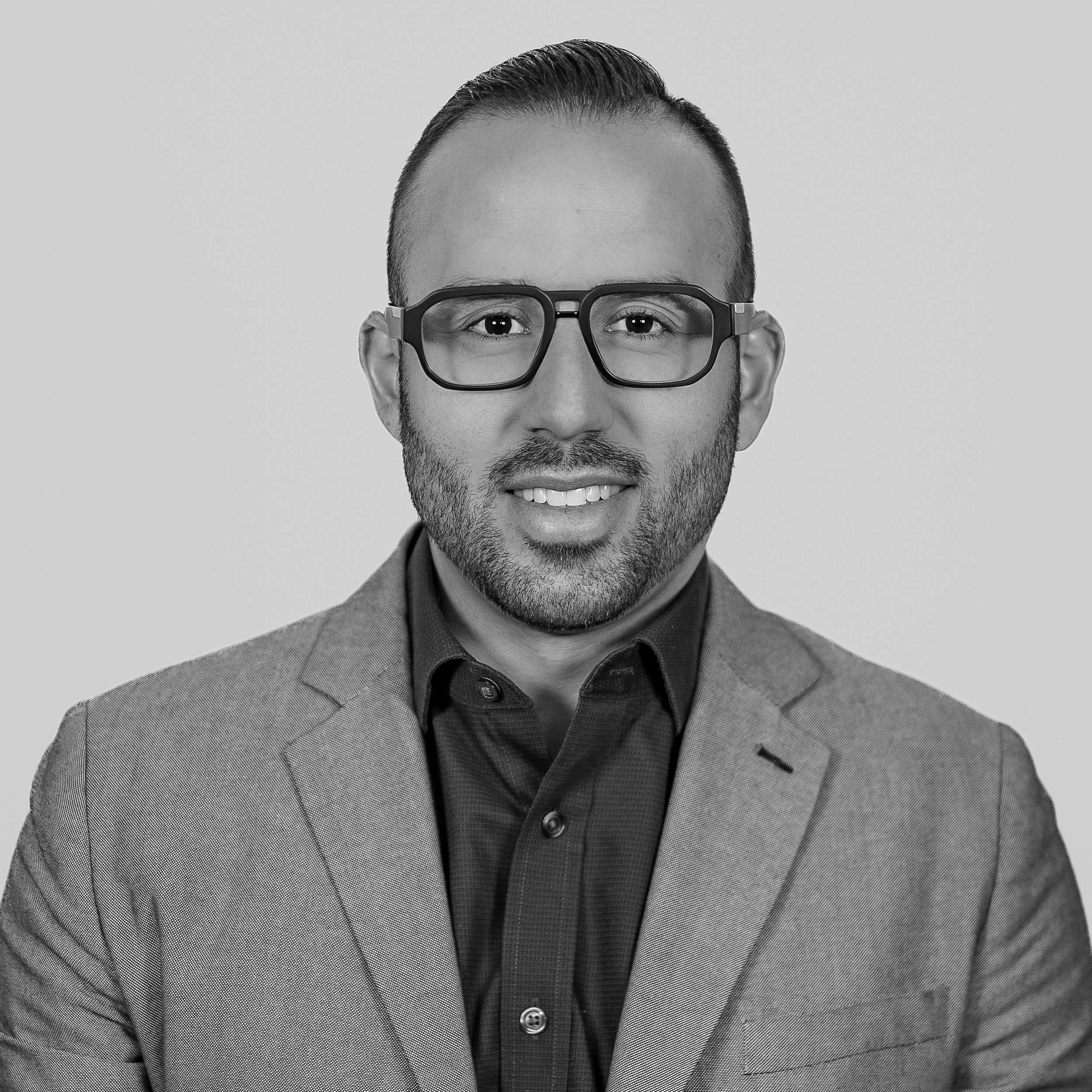 Black and white professional headshot of a man wearing glasses, a dark shirt, and a light-colored suit jacket, smiling at the camera.