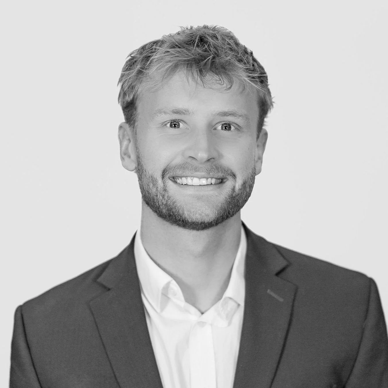 Black and white professional headshot of a smiling man with light-colored hair, beard, and mustache, wearing a suit and white shirt, facing forward.