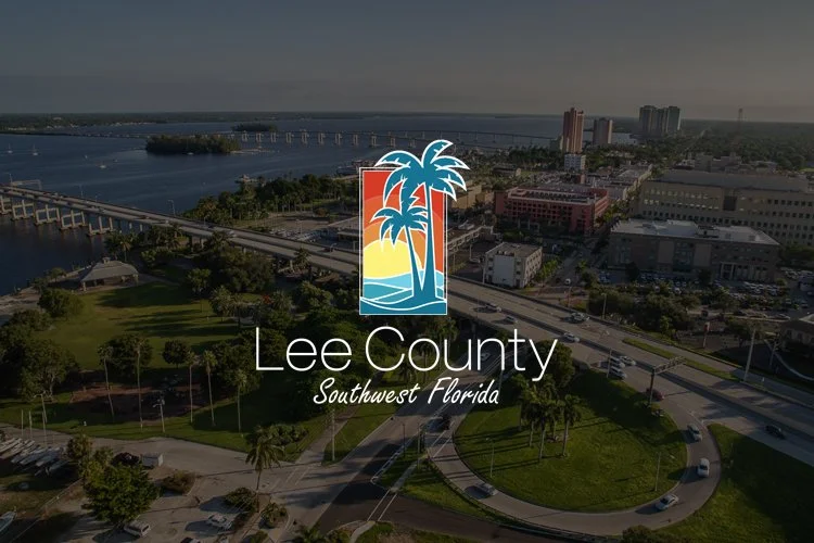Aerial view of Lee County, Southwest Florida, with a park, bridge, waterway, and city buildings, overlaid with the Lee County logo featuring palm trees and sunset colors.