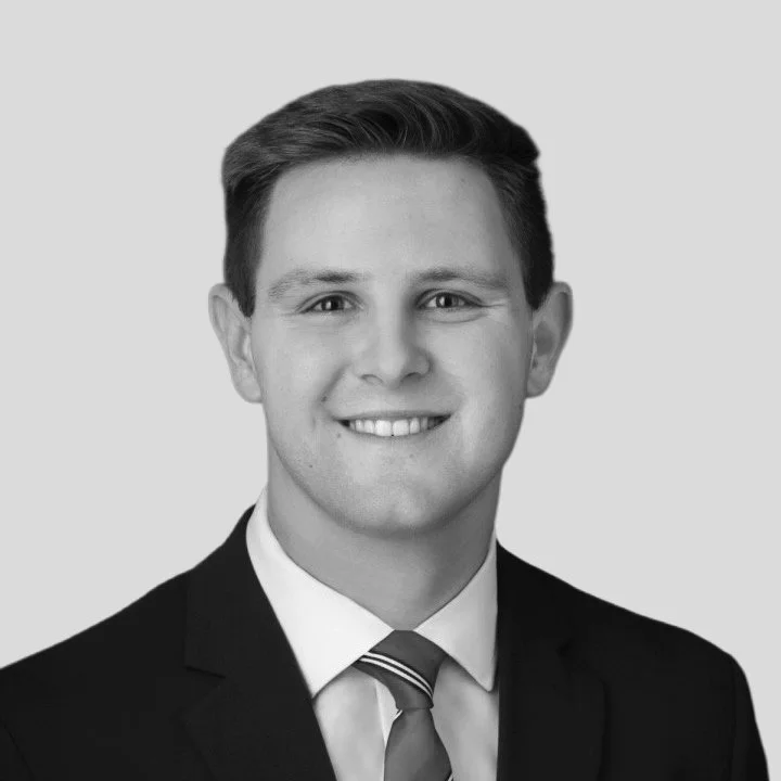 A black-and-white headshot of a young man with short, neatly styled hair, wearing a suit and tie, smiling slightly, against a plain background.