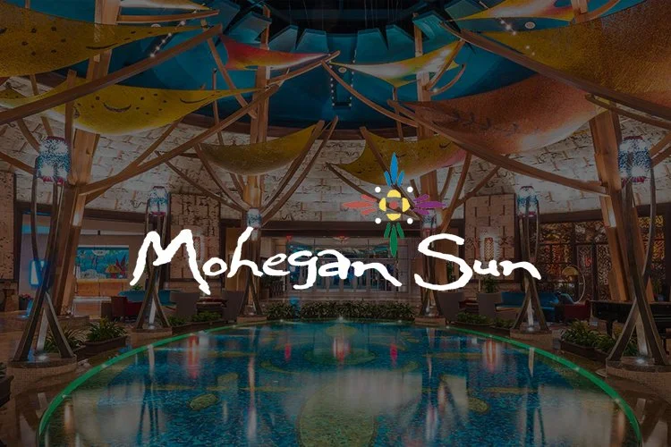 Indoor pool area with hanging fabric and wooden structures, decorated with colorful umbrellas and a Mohegan Sun logo.