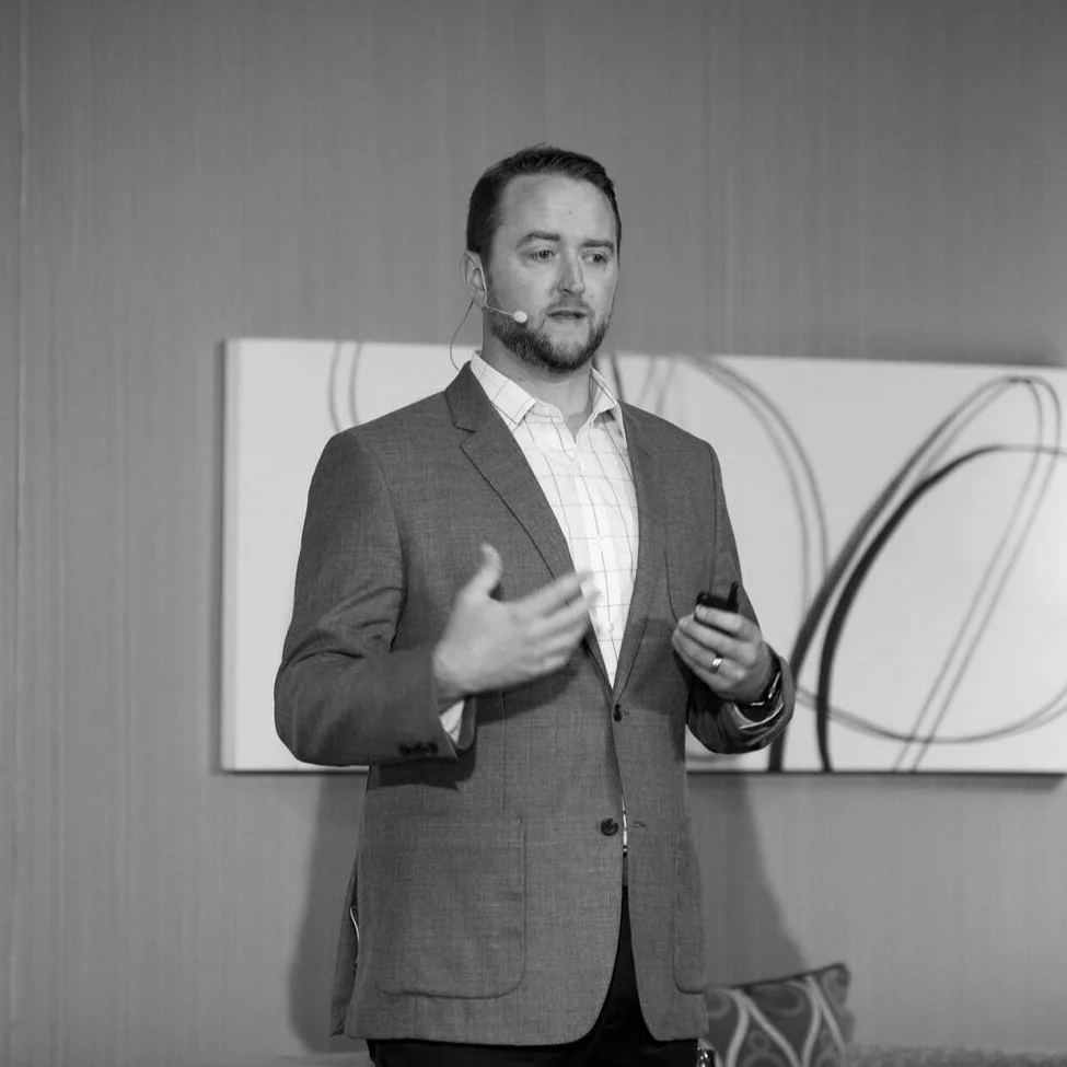 A man in a suit giving a presentation, wearing a headset microphone, and standing in front of abstract artwork on a wall.