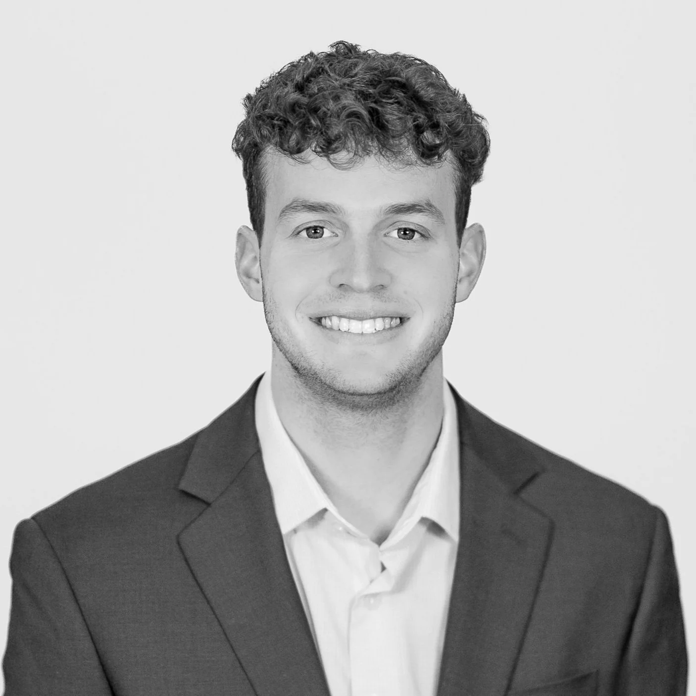 Black and white professional headshot of a young man with curly hair, wearing a suit and dress shirt, smiling against a plain background.