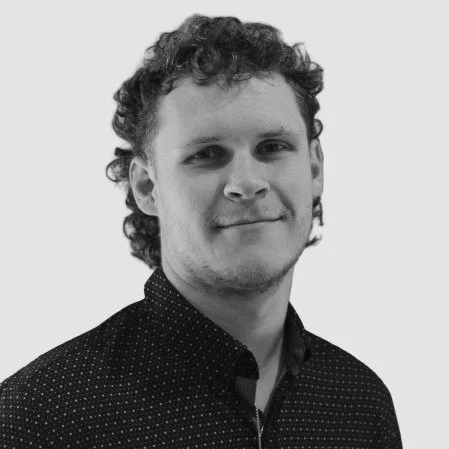 Portrait of a young man with curly hair, wearing a dark patterned shirt, against a light background.
