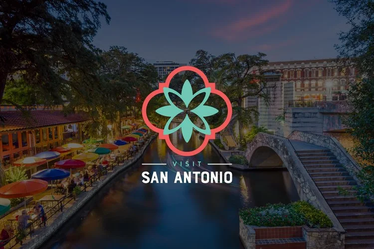 Nighttime view of downtown San Antonio with a river, colorful umbrellas on a walkway, and a bridge in the background, featuring an overlay of the San Antonio logo with a stylized flower and the text 'Visit San Antonio'.
