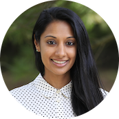 A young woman with long black hair, smiling, wearing a white polka dot blouse, outdoors with greenery in the background.